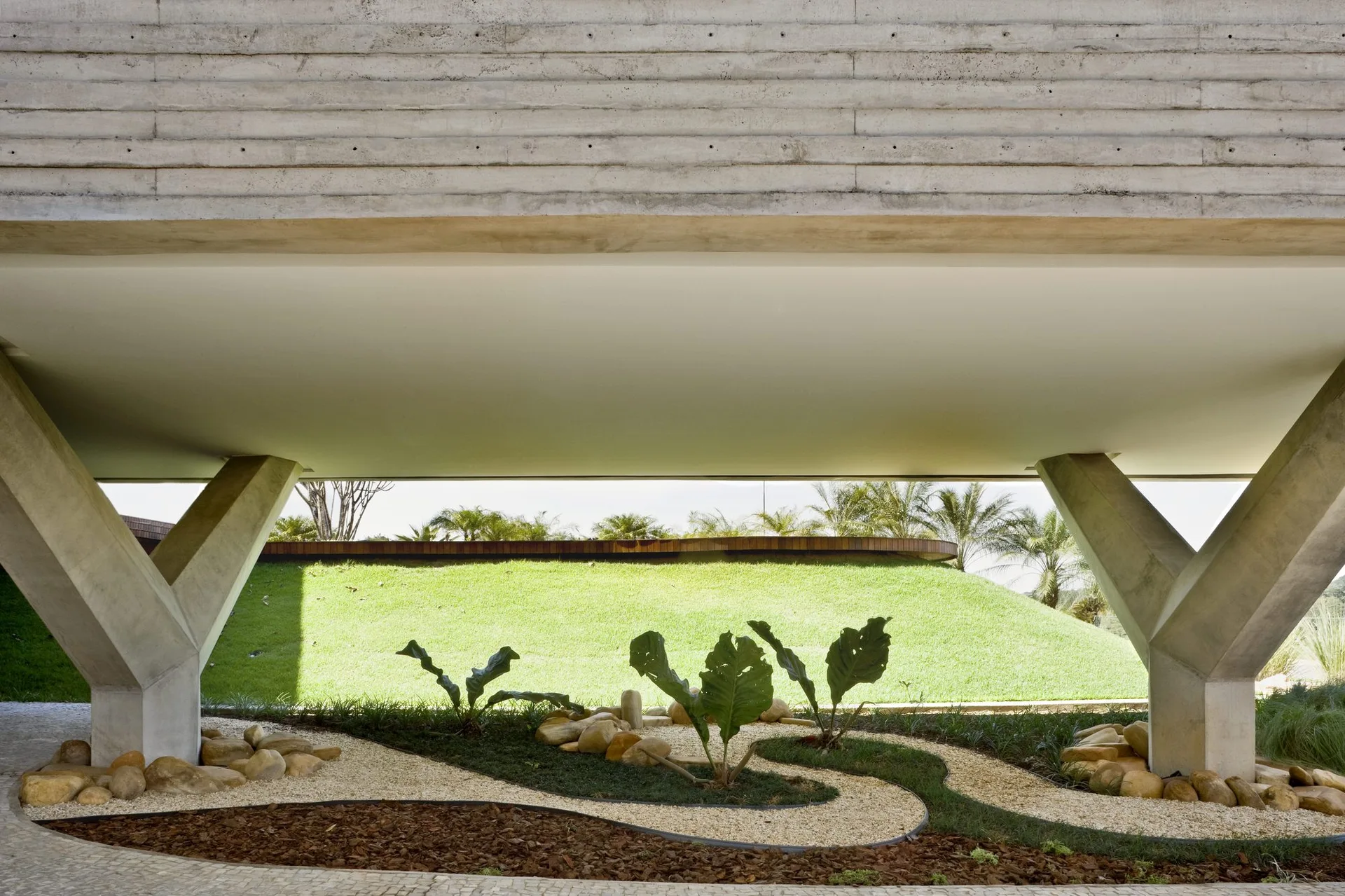 Angular concrete columns support a massive horizontal beam, framing a landscaped garden with curved pebble paths, rocks, and green foliage.