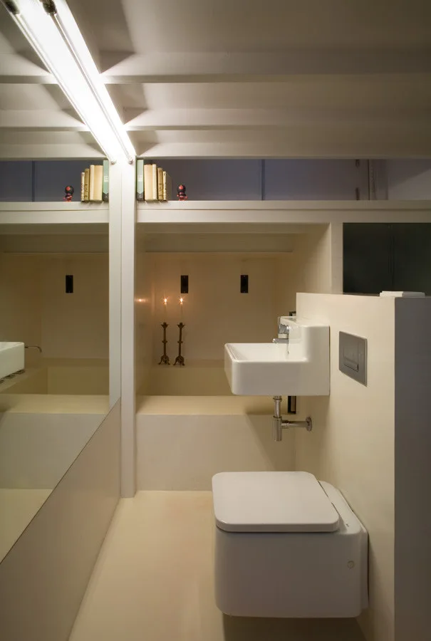 A minimalist off-white bathroom features a wall-hung toilet, a floating sink, exposed ceiling beams, and a large mirror that reflects the space.