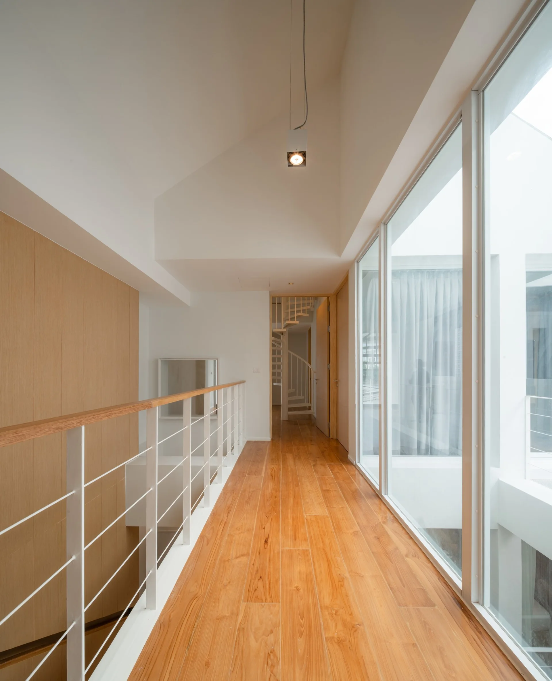 A modern hallway features a light wood paneled wall, warm wood flooring, a white metal railing, and a wall of large glass windows.