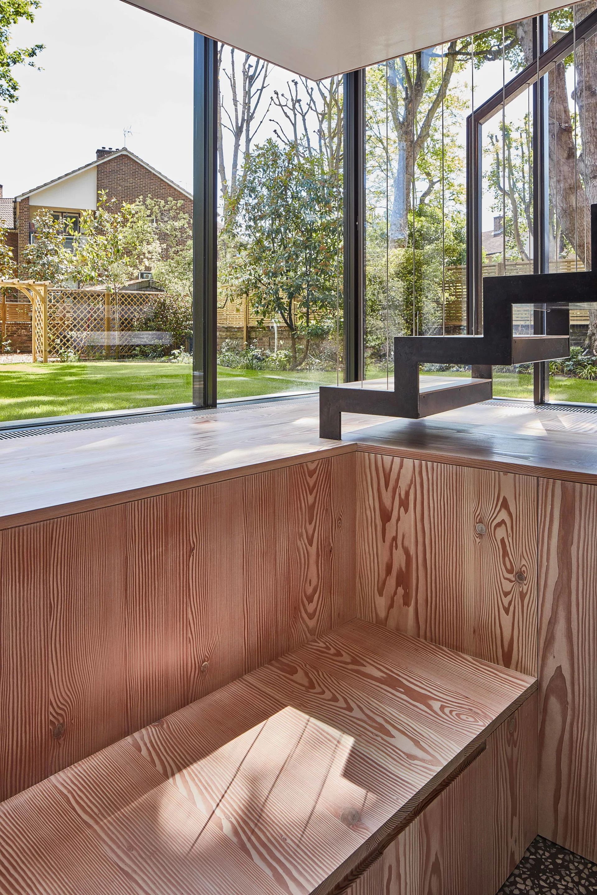A black metal open-riser staircase stands above natural wood cabinets next to a floor-to-ceiling glass wall looking onto a green lawn.
