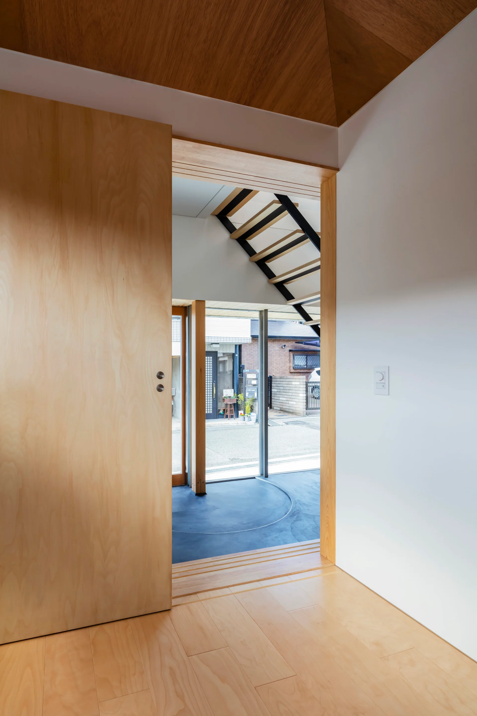 A minimalist entryway features a large light wood sliding door, matching wood flooring, and an open riser staircase visible in the background.
