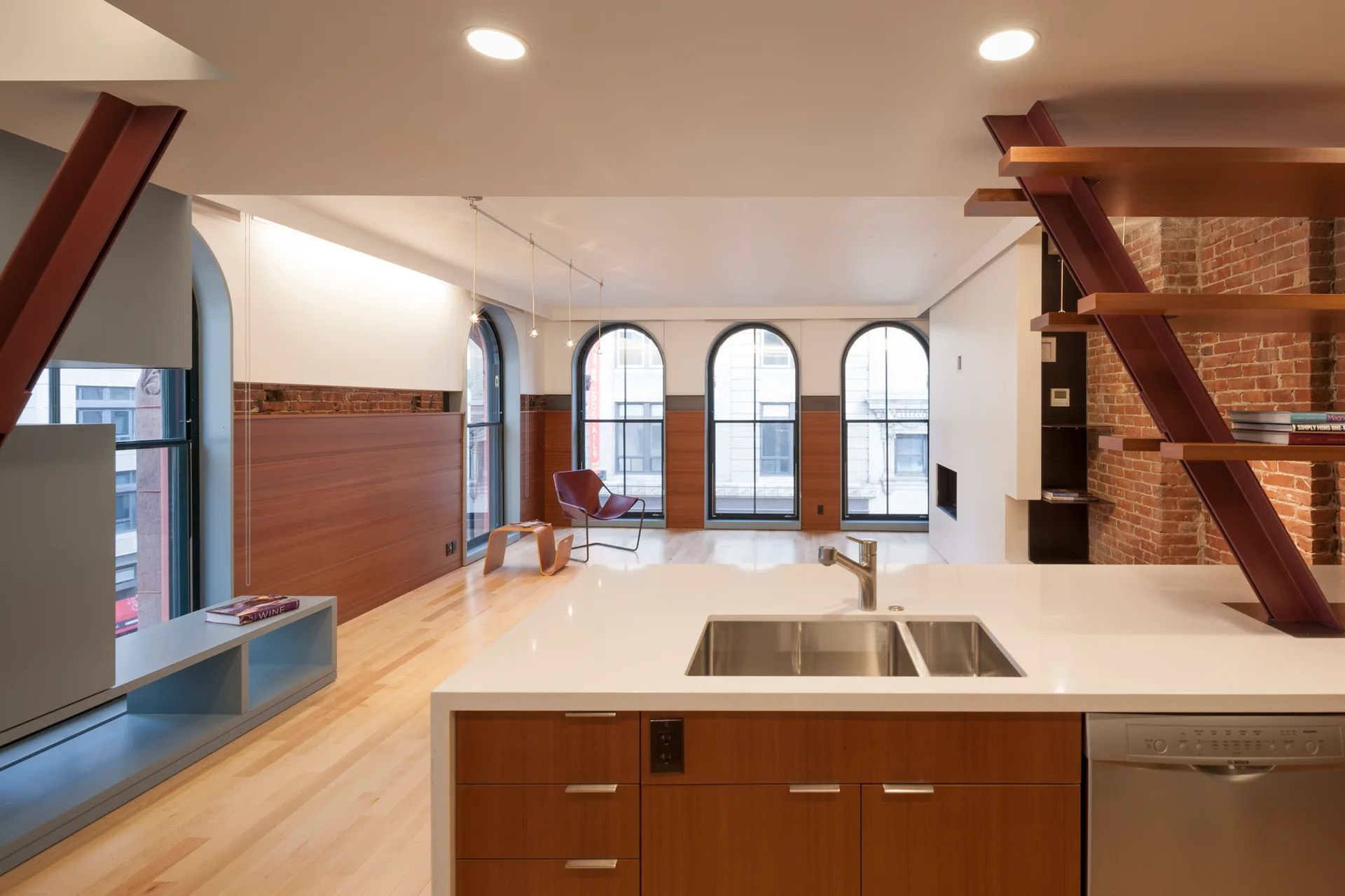An open-plan kitchen island with a white countertop faces a living area featuring arched windows, wood-paneled walls, and a metal staircase.