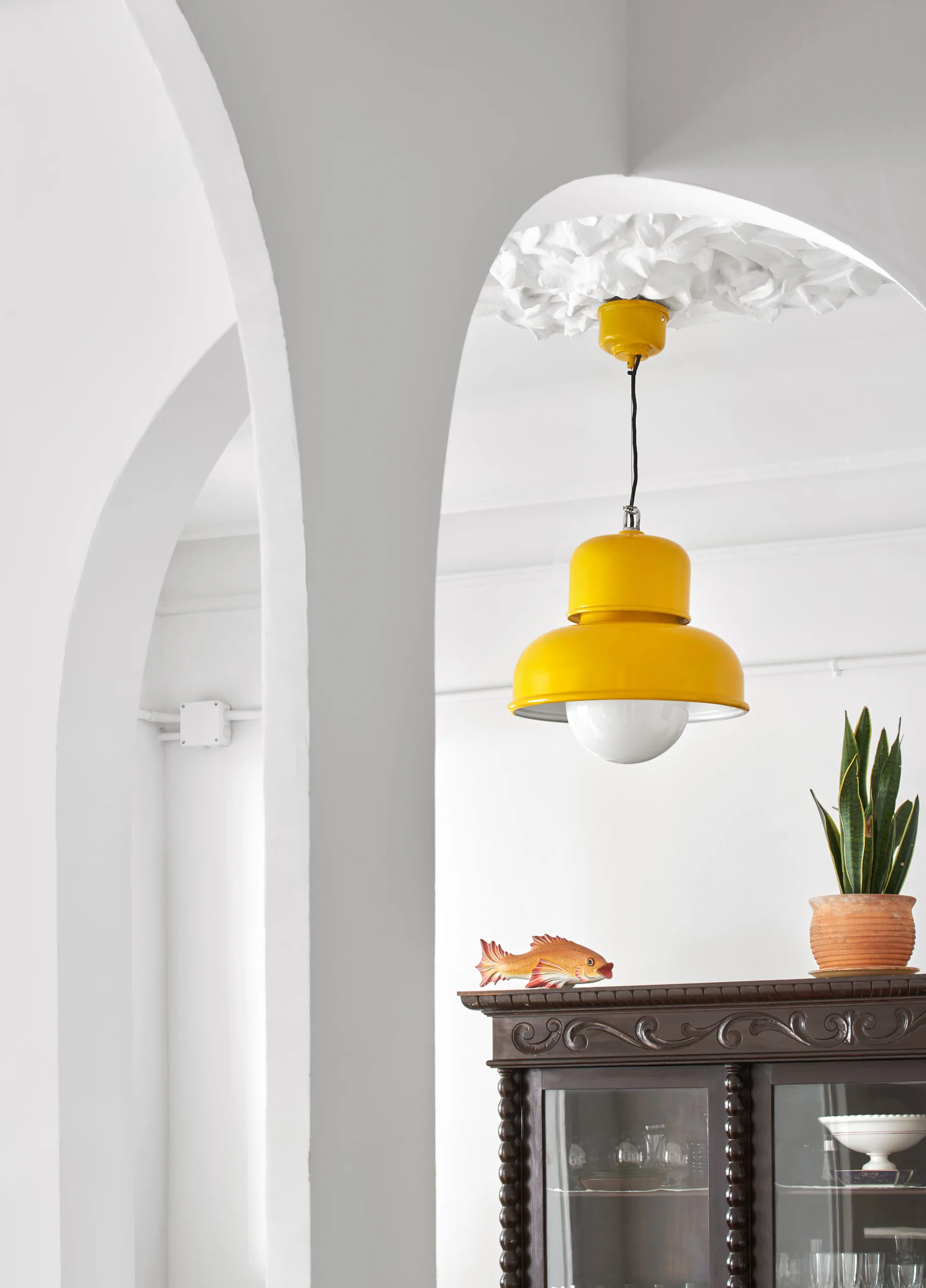 A bright yellow pendant light hangs from an ornate ceiling medallion behind a white plaster archway, above a dark wood glass-front cabinet.