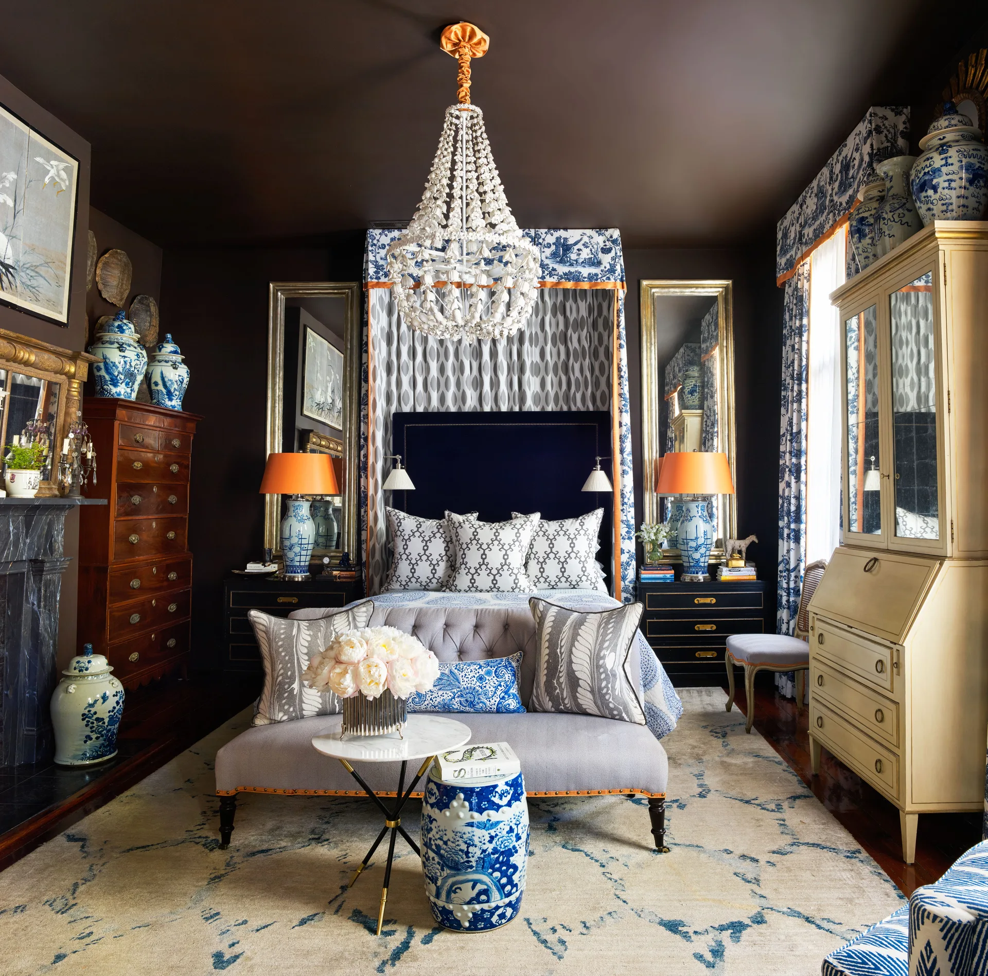 A maximalist bedroom with dark brown walls, a fabric canopy bed, a shell chandelier, blue and white porcelain accents, and a gray tufted bench.