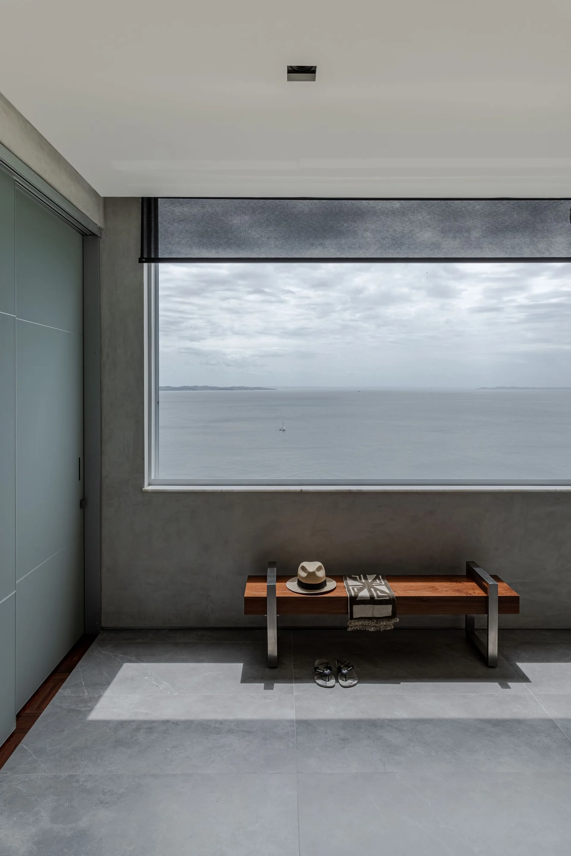 A low wooden bench with a hat sits on a gray tiled floor in front of a large window overlooking the ocean.