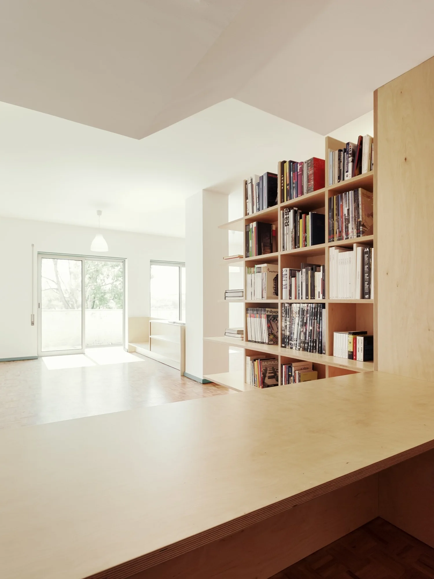 A light-wood plywood bookshelf stands next to a custom desk in a bright room with parquet floors and sliding glass doors.
