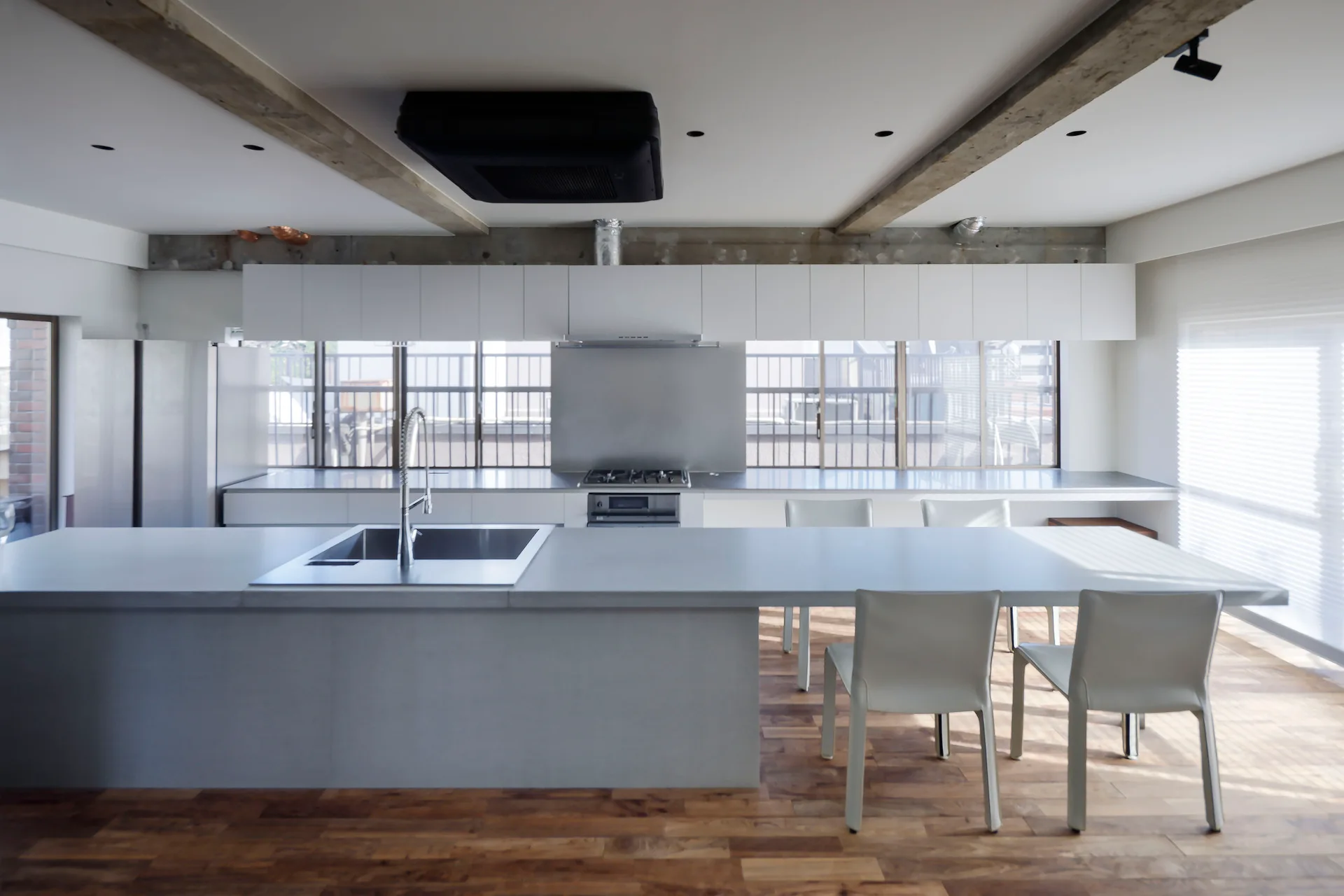 A minimalist kitchen features a long grey island with a sink, white upper cabinets above a ribbon window, and exposed ceiling beams.