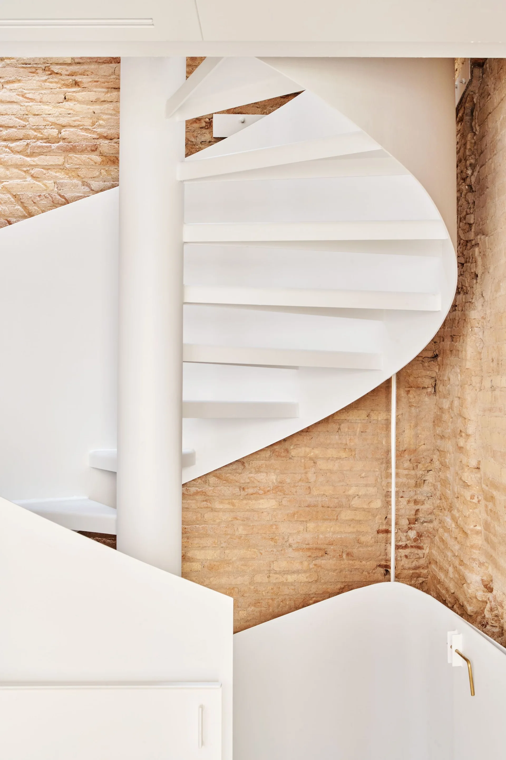 A white metal spiral staircase winds upward in front of a textured, tan exposed brick wall.