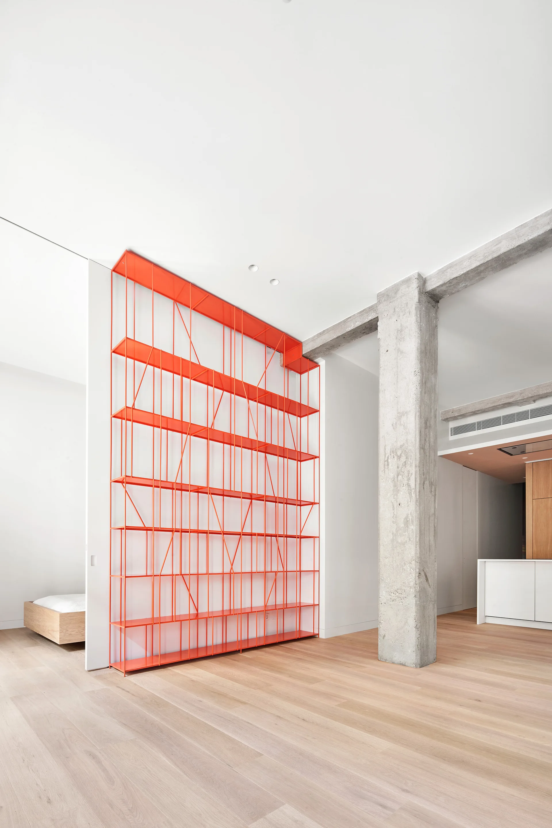 A bright minimalist room features a tall orange metal shelving unit next to an exposed concrete column and light wood floors.