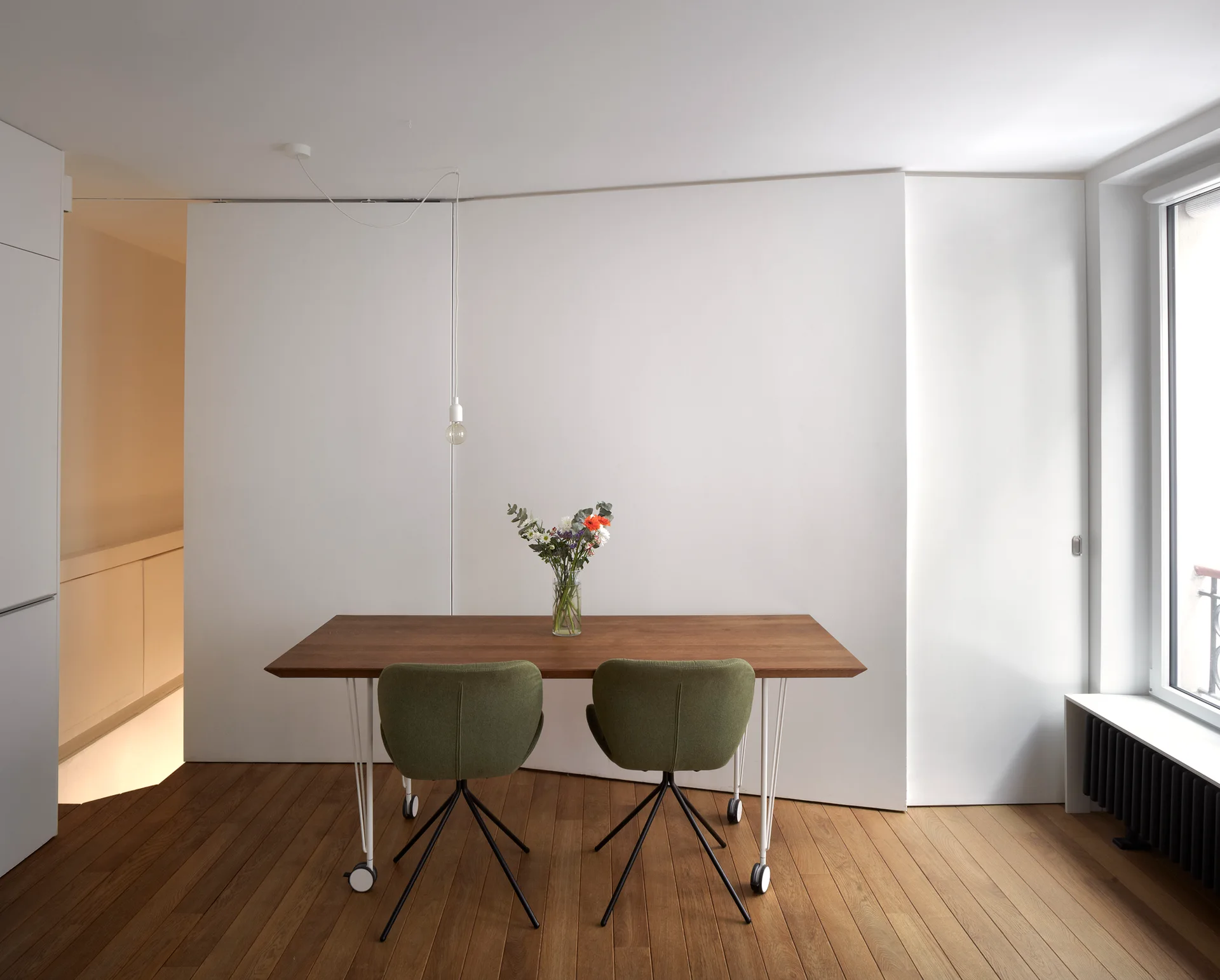 A wooden dining table paired with two olive green chairs sits on a hardwood floor against a clean white wall with a hanging lightbulb.