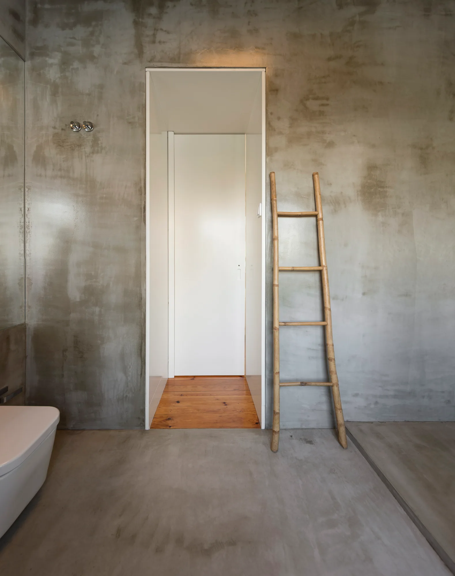 A minimalist gray concrete bathroom features a wooden ladder leaning against the wall next to an open doorway showing wood floors.