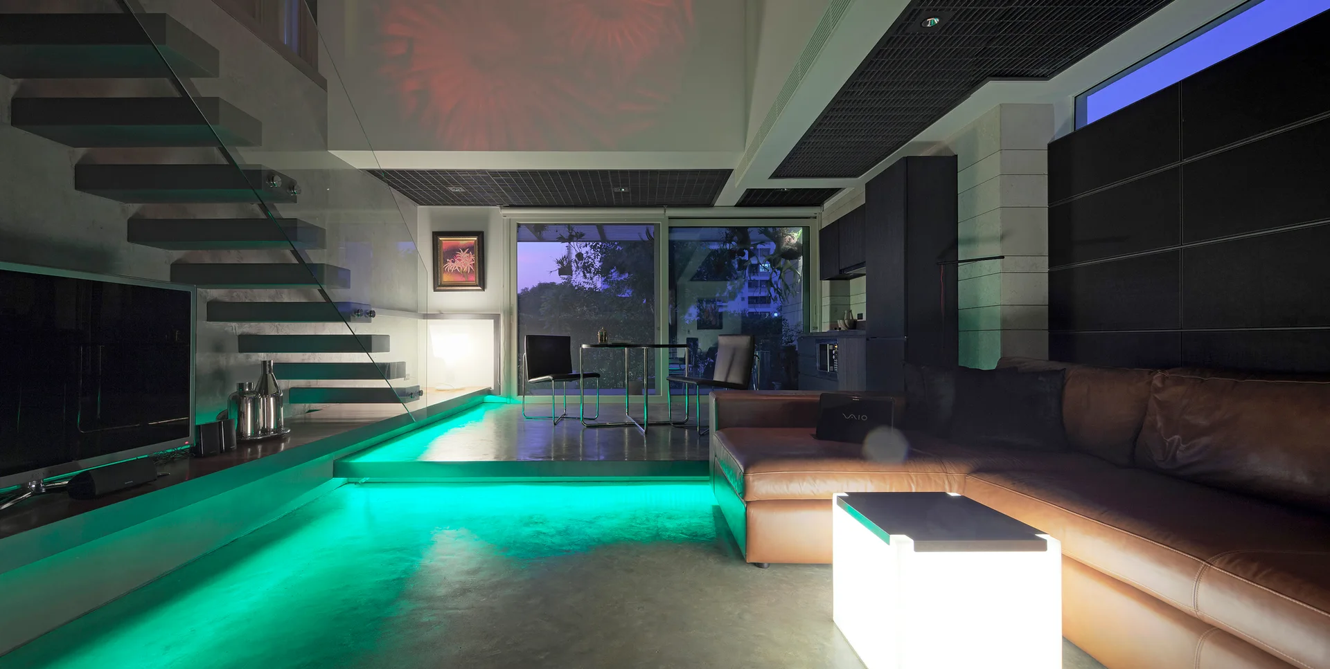 A modern living room features a brown leather sectional, a floating staircase with glass railing, and green LED accent lighting under the platform.