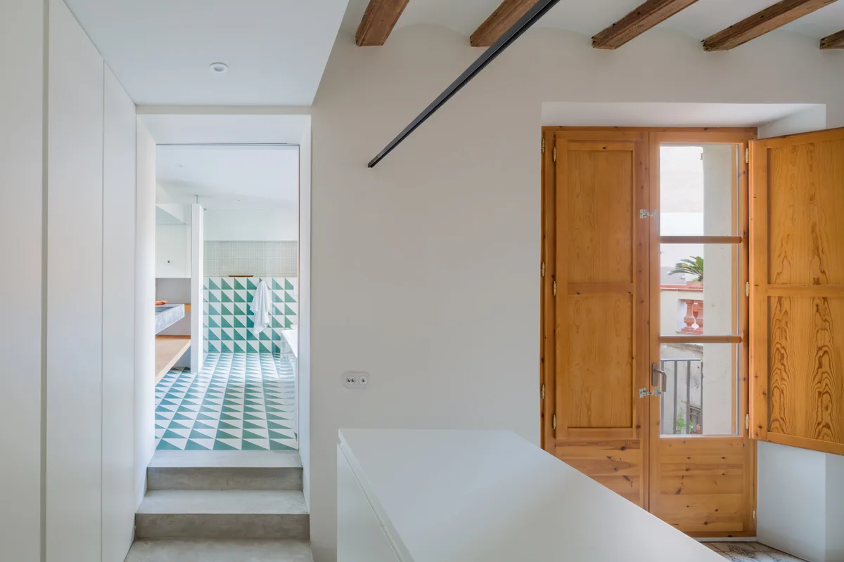 A white hallway with exposed wooden ceiling beams and double doors features concrete steps leading into a bathroom with green and white geometric floor tiles.