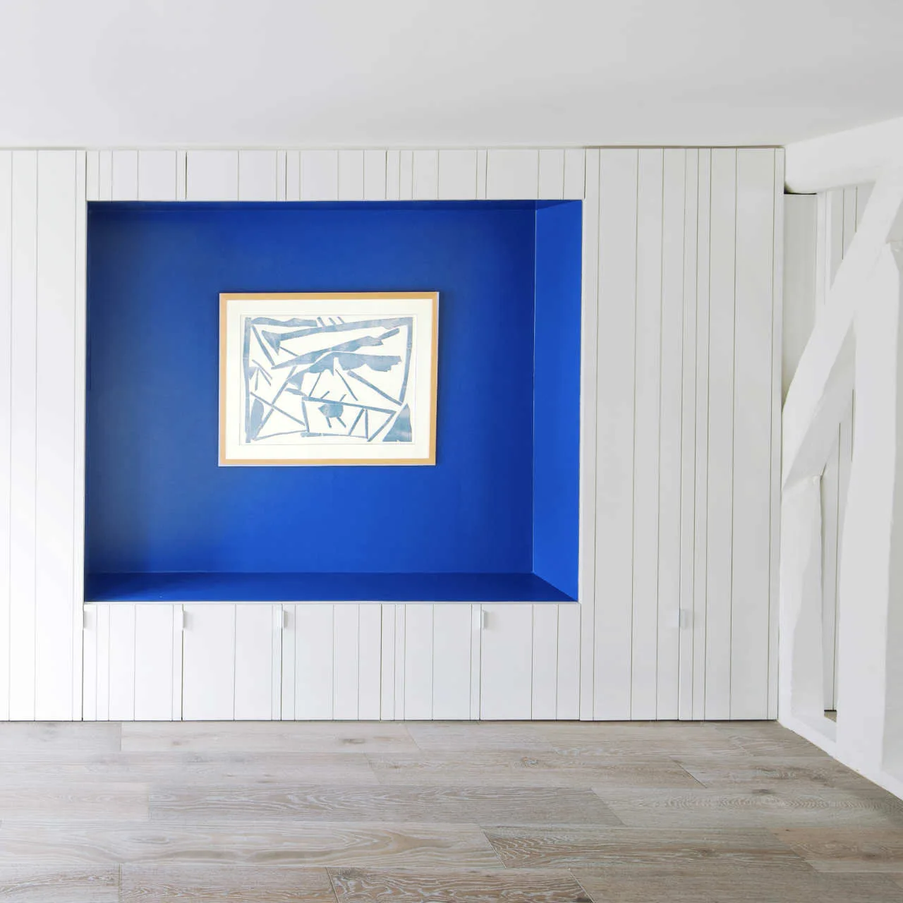 A white paneled wall with a large, deep-blue rectangular alcove containing a framed piece of abstract art above light wood flooring.