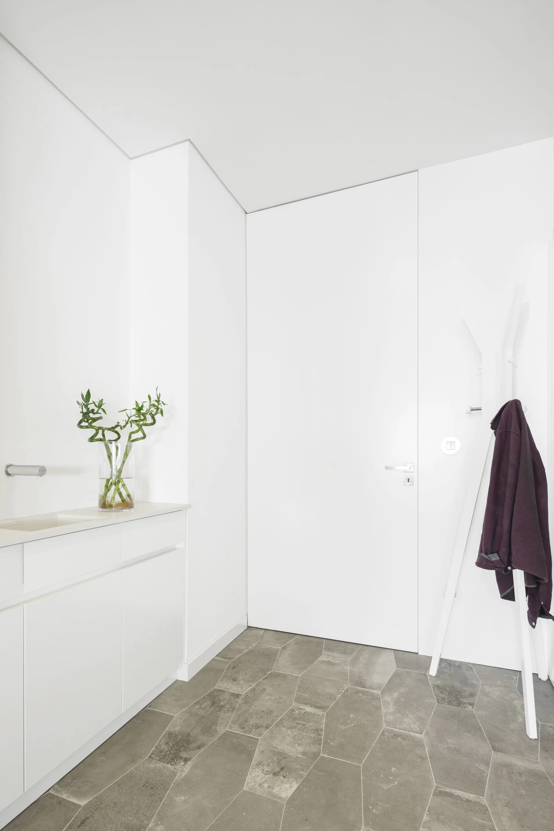 A minimalist white entryway featuring a flush door, a white built-in cabinet with a plant, a coat rack, and grey geometric stone floor tiles.