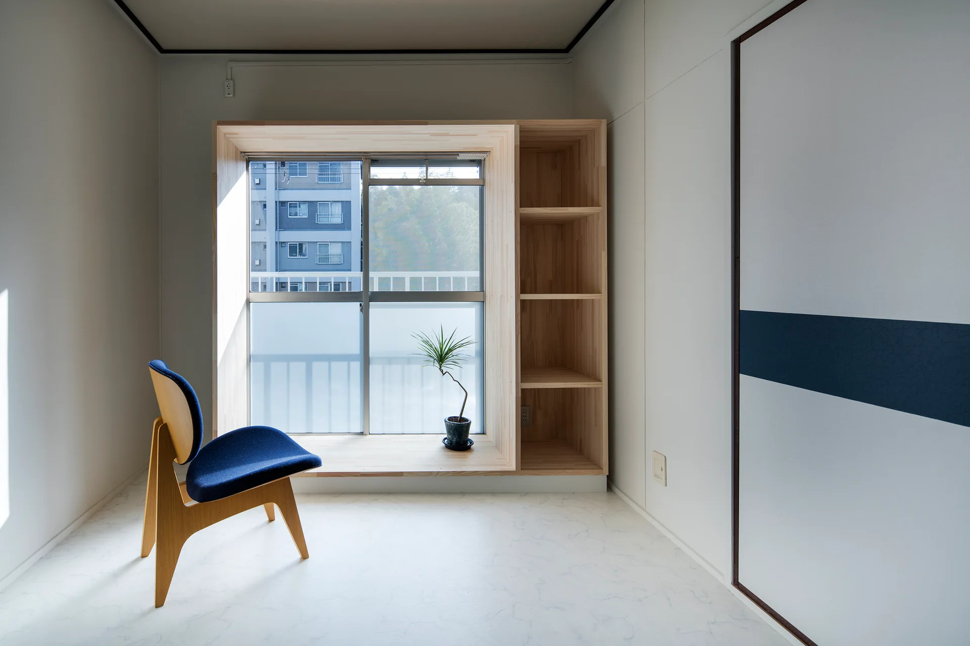 A minimalist room features a blue upholstered accent chair next to a custom light wood window seat with integrated vertical shelving.