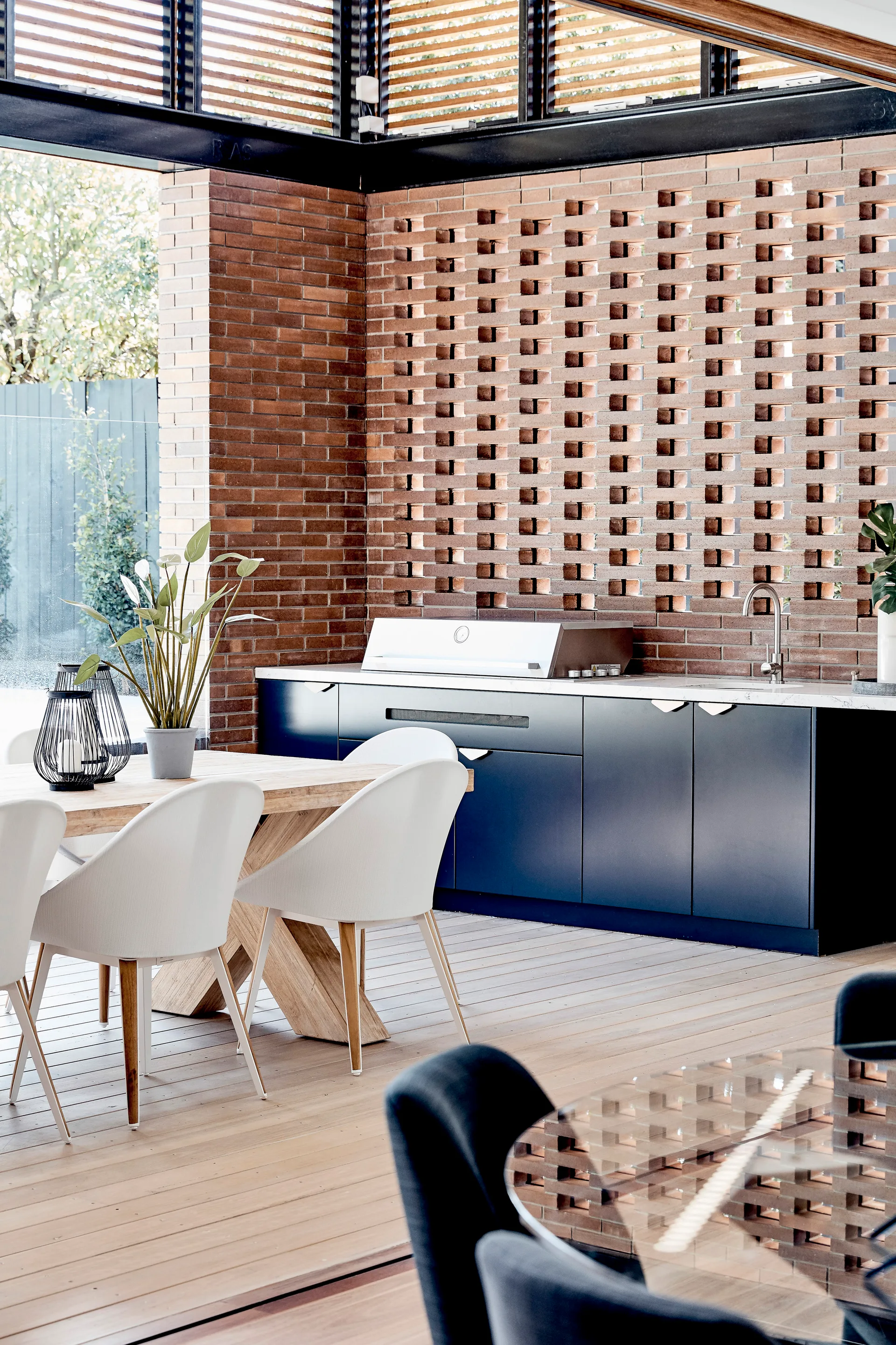 An outdoor kitchen with dark cabinetry and a dining table is set against a perforated red brick wall on a wooden deck.