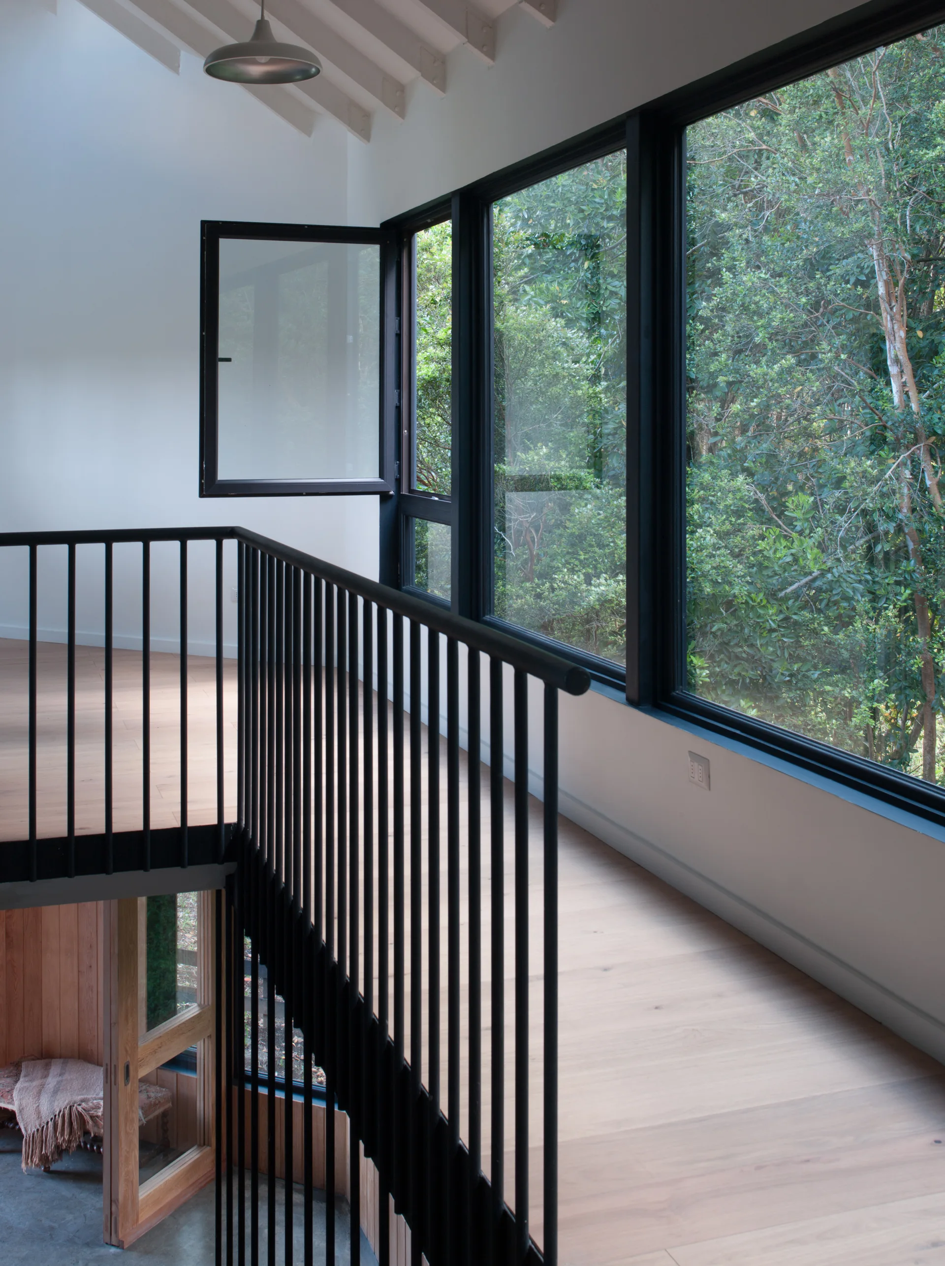 A modern home landing features light wood floors, a black metal spindle railing, and large black-framed windows overlooking a lush green forest.