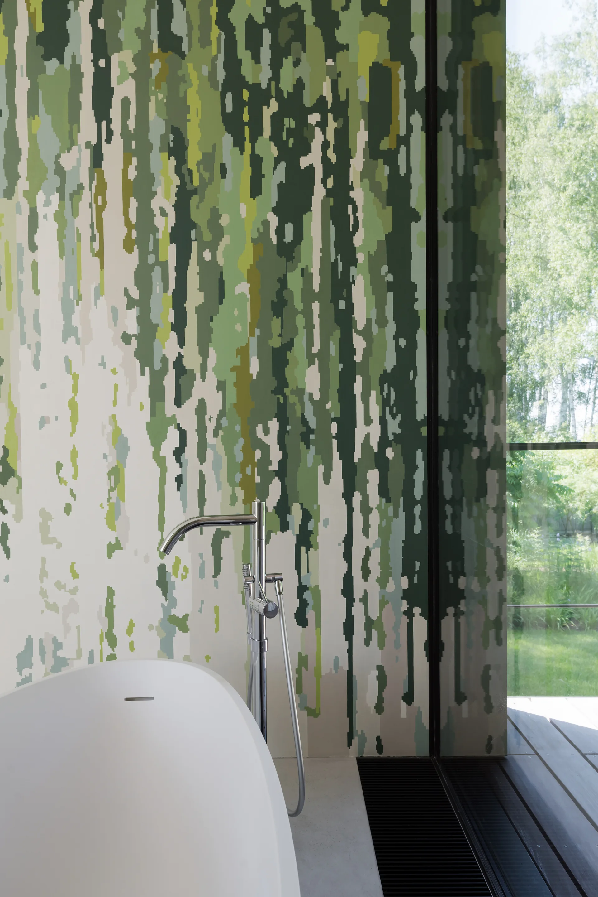 A white freestanding bathtub and chrome floor faucet stand in front of an abstract green and white patterned wall near a large window.