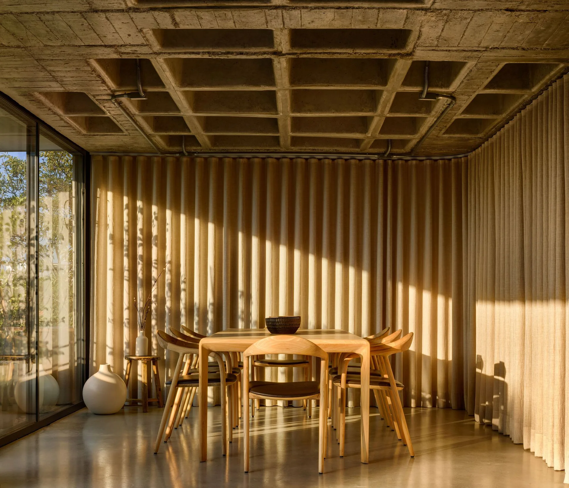 A wooden dining set stands on a polished concrete floor beneath an exposed concrete waffle ceiling, framed by floor-to-ceiling pleated beige curtains.