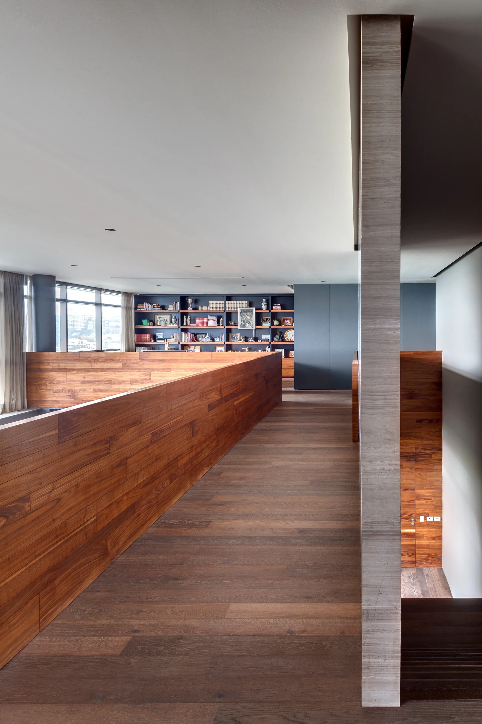 A modern hallway landing featuring a wide wood-clad partition wall, a veined stone pillar, and a built-in bookshelf in the background.