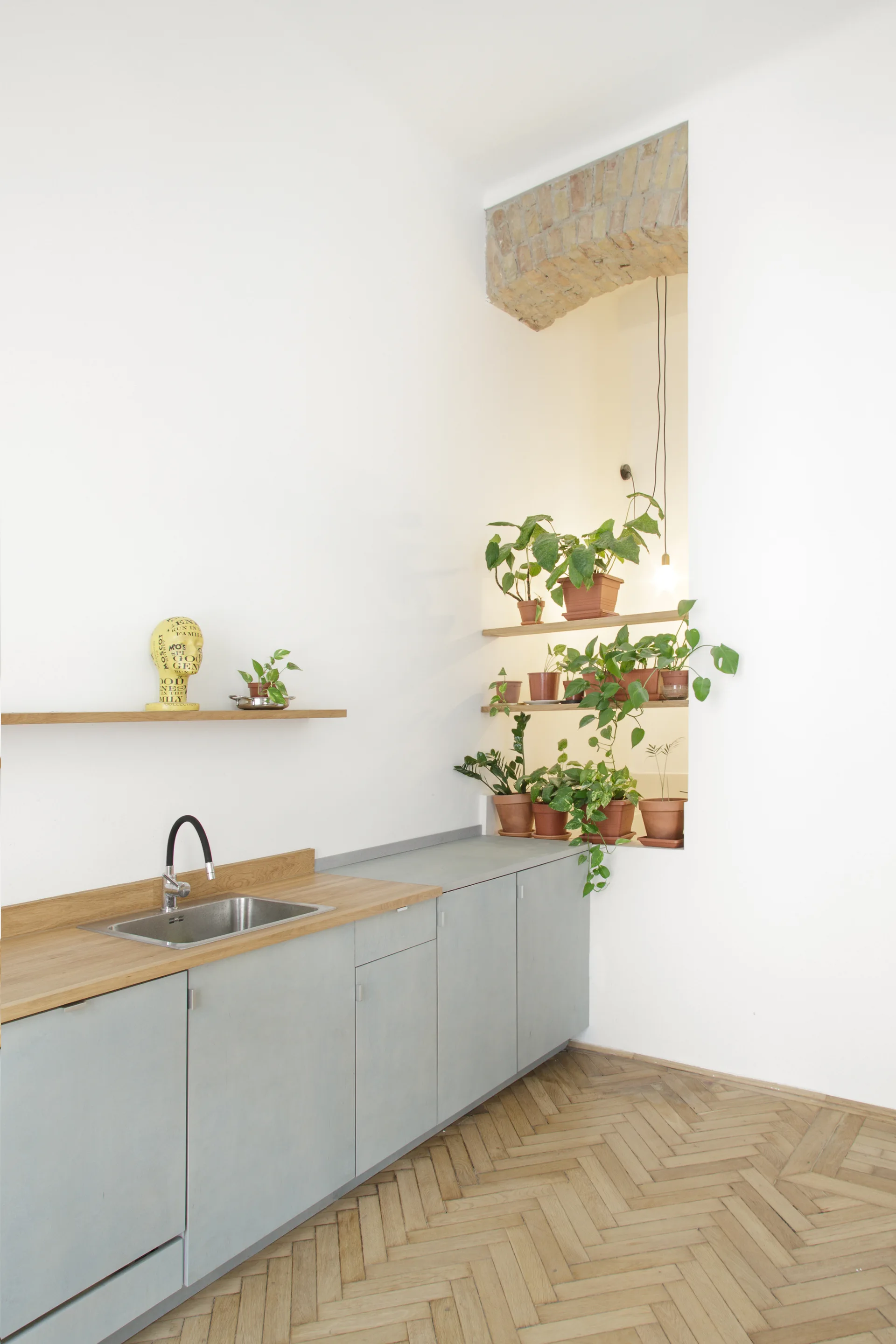 Minimalist grey kitchen cabinets with a wood countertop stand on a herringbone floor next to a tall wall niche filled with potted plants.