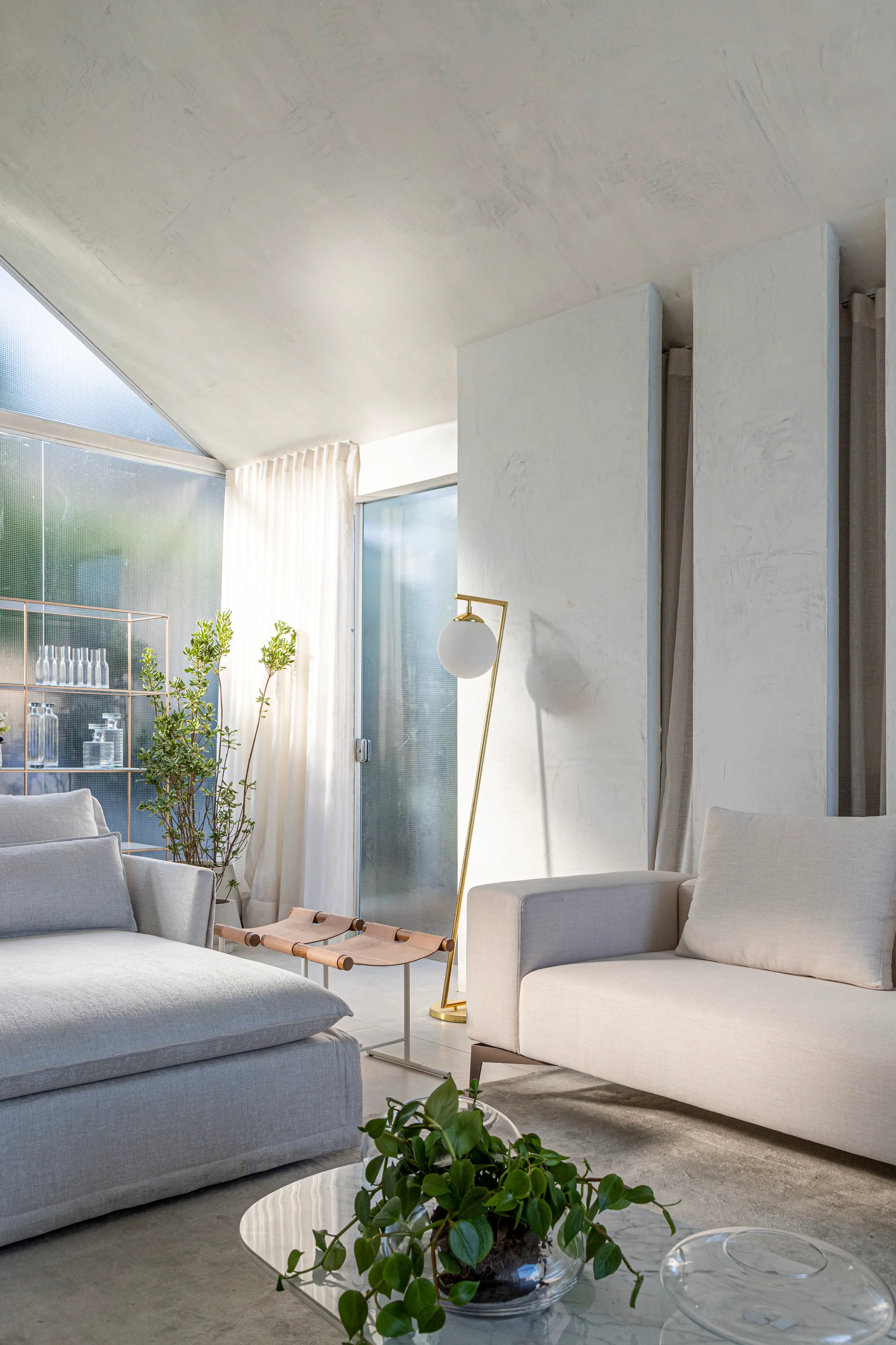 A contemporary living room featuring light beige upholstered sofas, a slender gold floor lamp, textured white plaster walls, and a marble coffee table.