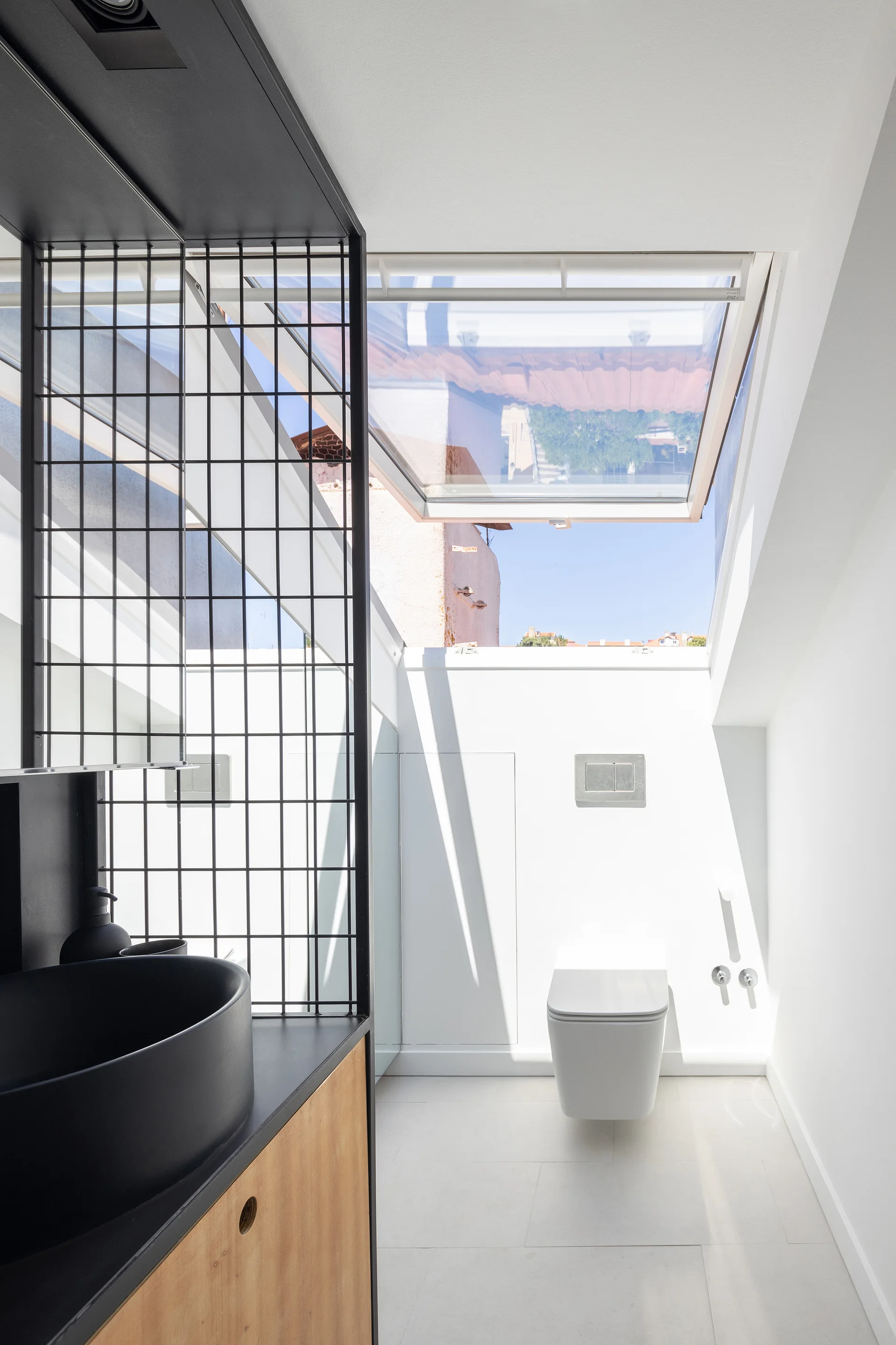 A modern white bathroom featuring a wall-mounted toilet, an open skylight in the ceiling, and a black vessel sink with a black metal grid partition.