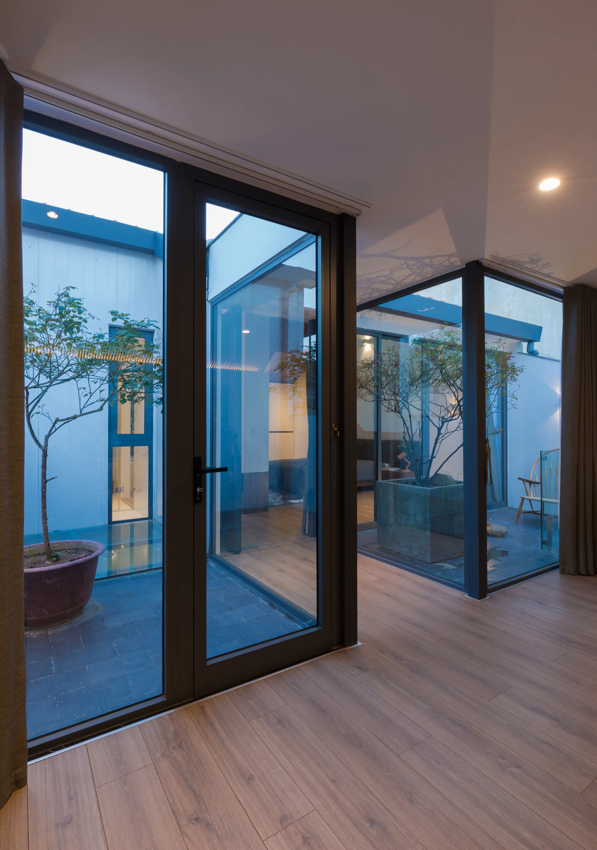 A modern interior with light wood flooring faces an enclosed courtyard with glass doors, containing stone planters and small trees.