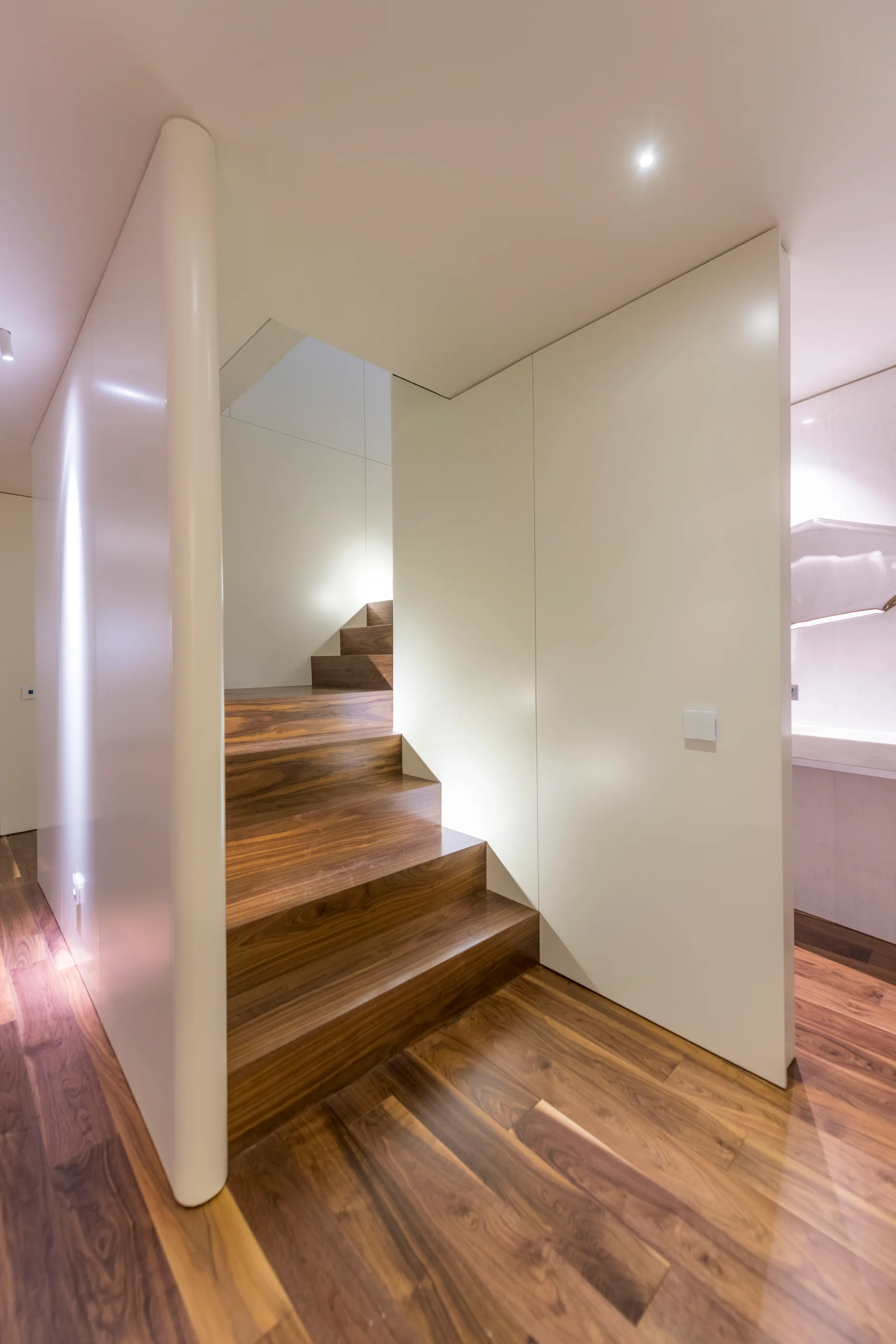 A minimalist modern staircase with rich walnut steps rising between smooth, off-white wall panels.