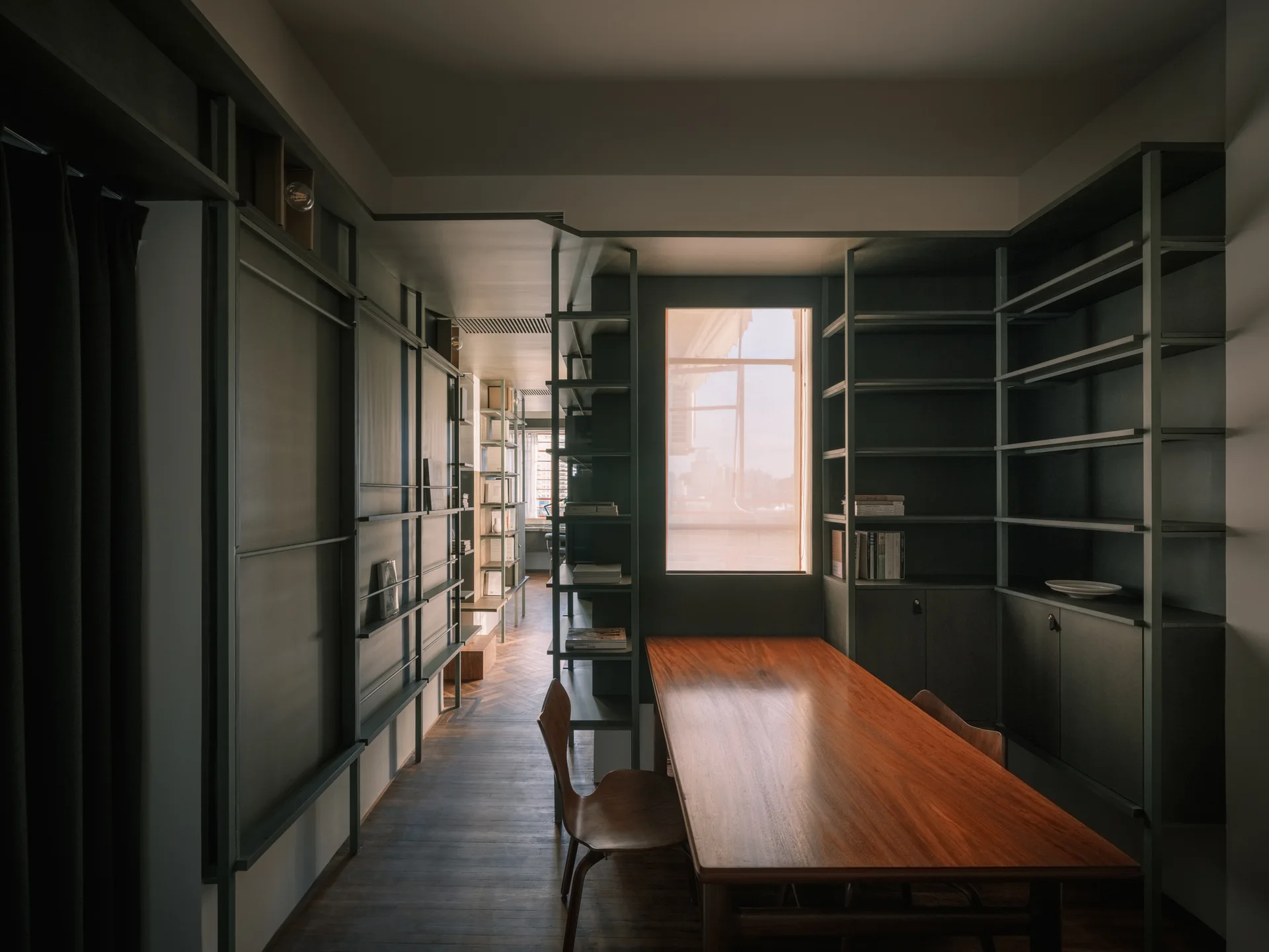 A home library features matte dark green shelving on the walls, a long polished wood table with a molded wood chair, and herringbone hardwood floors.