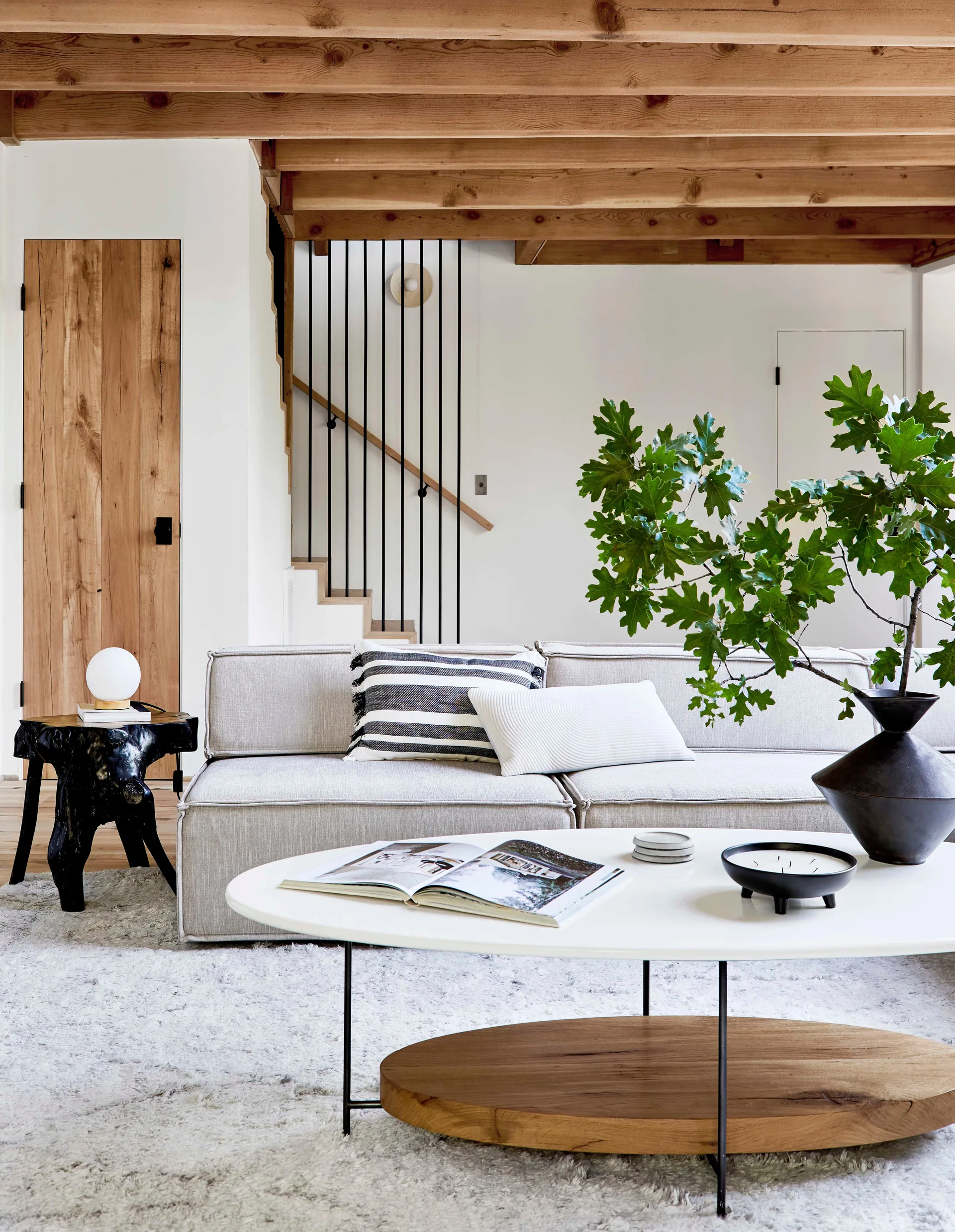 A light grey sofa is paired with a white oval coffee table on a plush shag rug beneath exposed natural wood ceiling beams.