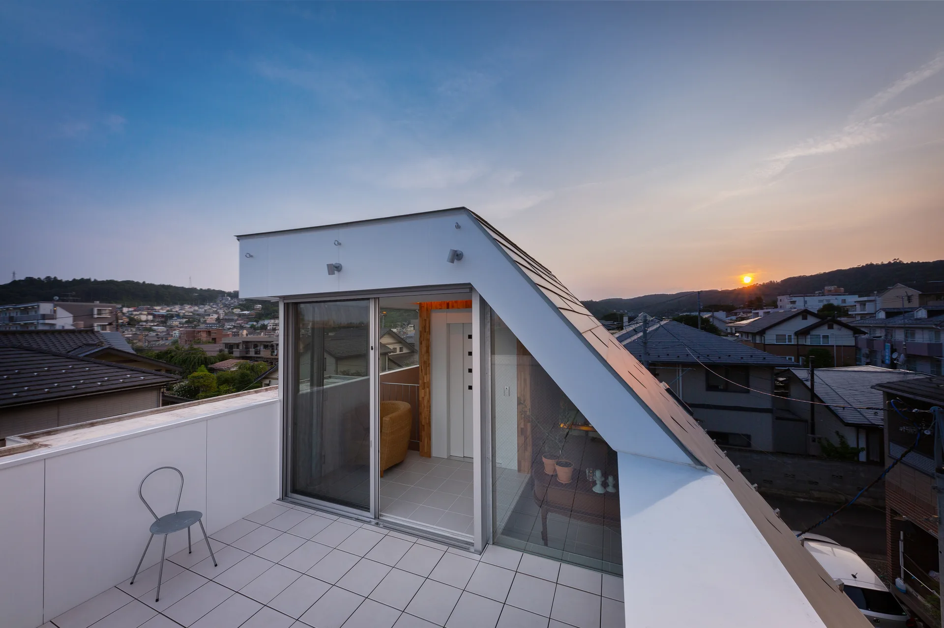 A modern rooftop terrace features white tiled flooring, a single metal chair, and a sloped architectural structure with sliding glass doors at sunset.