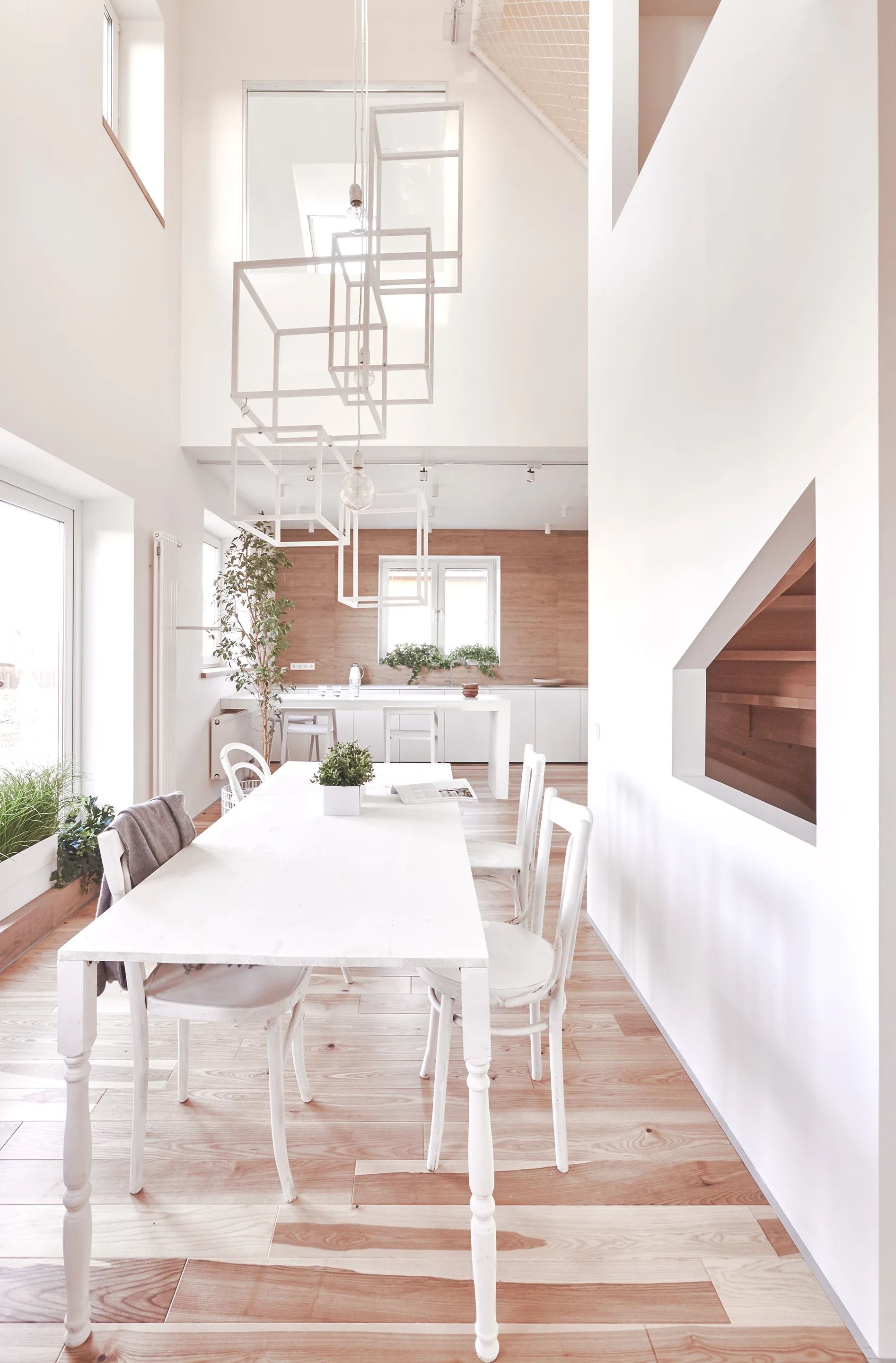 A bright double-height dining room features a white wooden table and chairs, light wood flooring, and an open geometric cube pendant light fixture.
