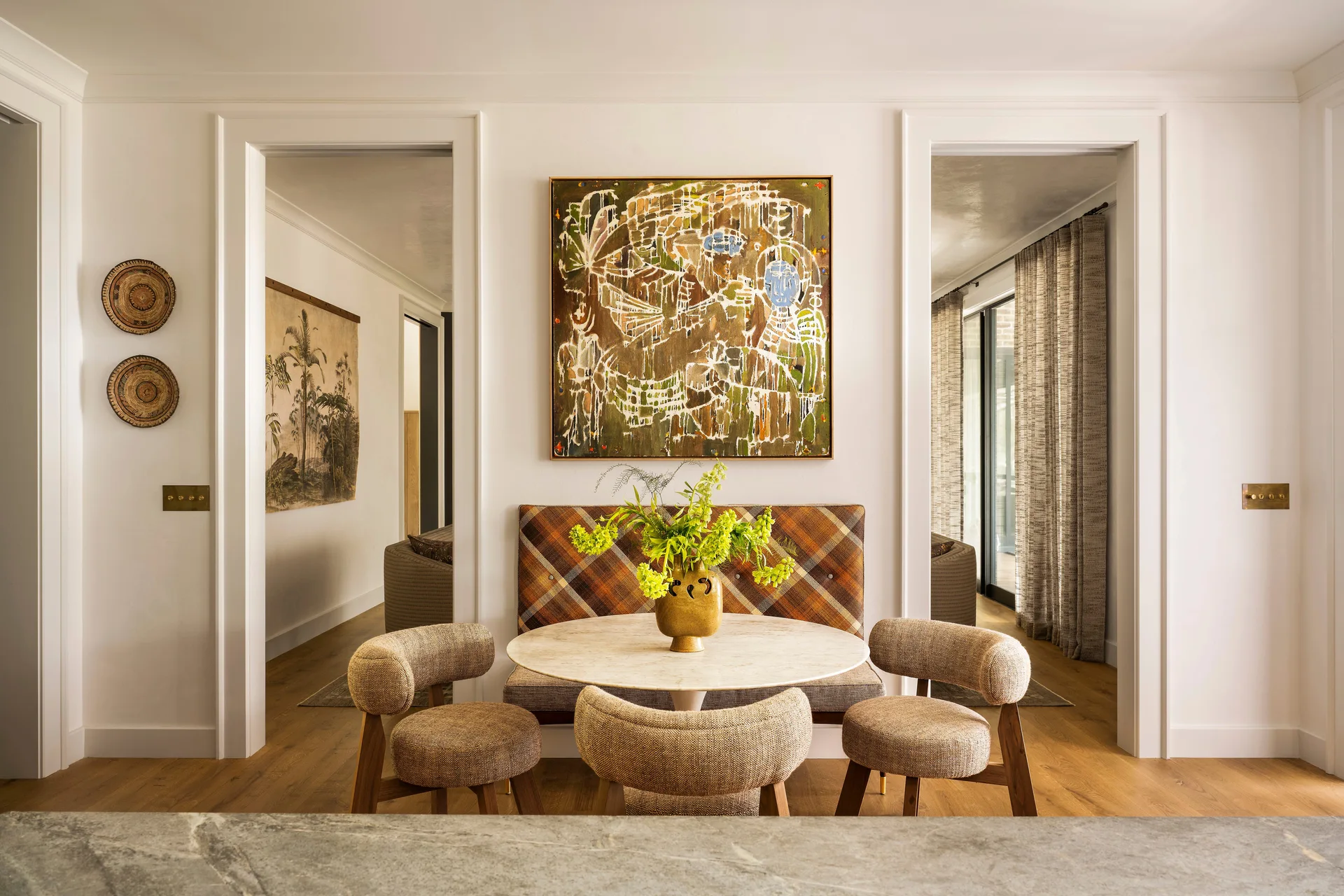 A dining nook features a round marble table, three textured upholstered chairs, and a plaid banquette under a large abstract painting between two doorways.