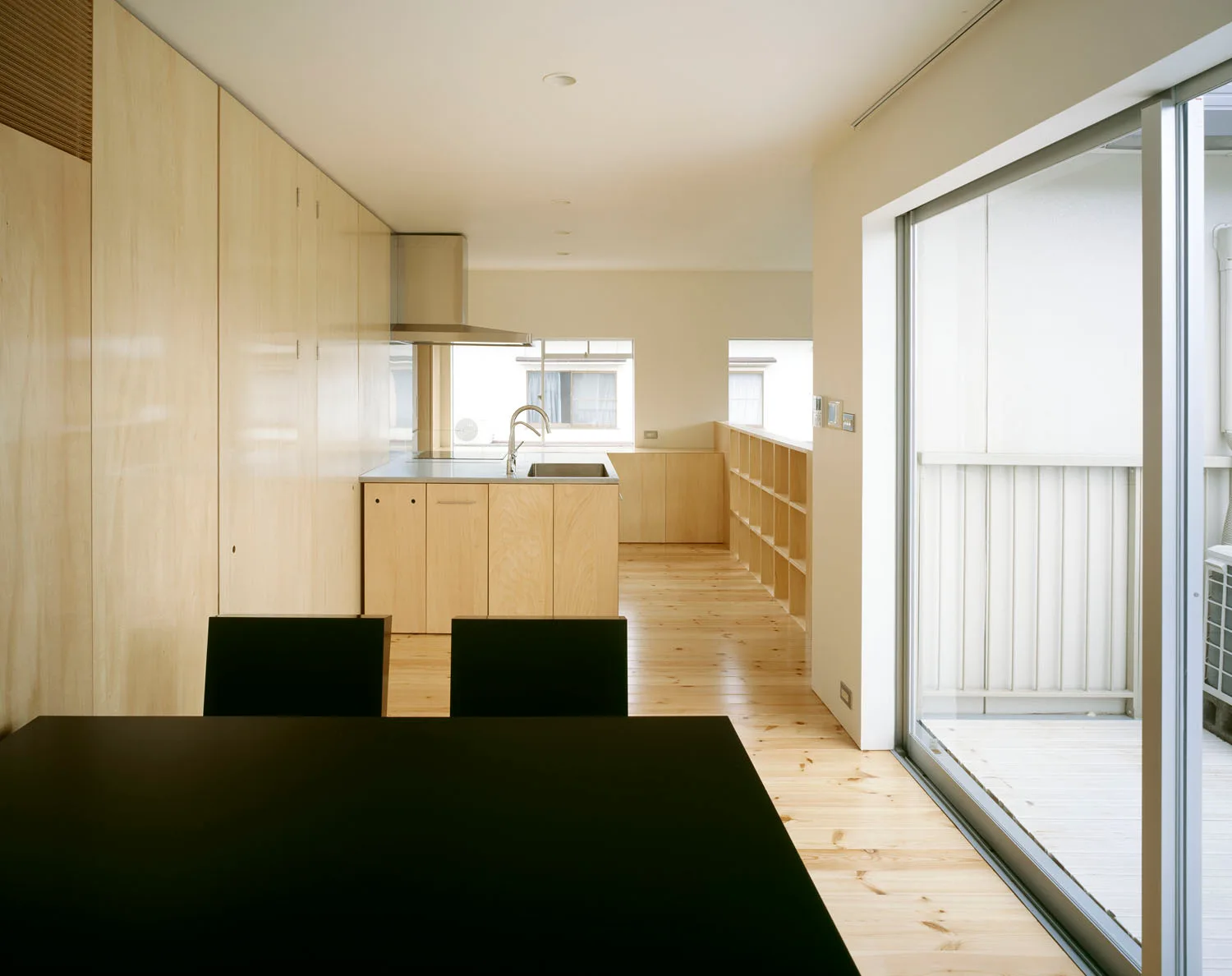 A minimalist kitchen features light wood plywood cabinetry, matching wood floors, a stainless steel range hood, and a dark dining table in the foreground.