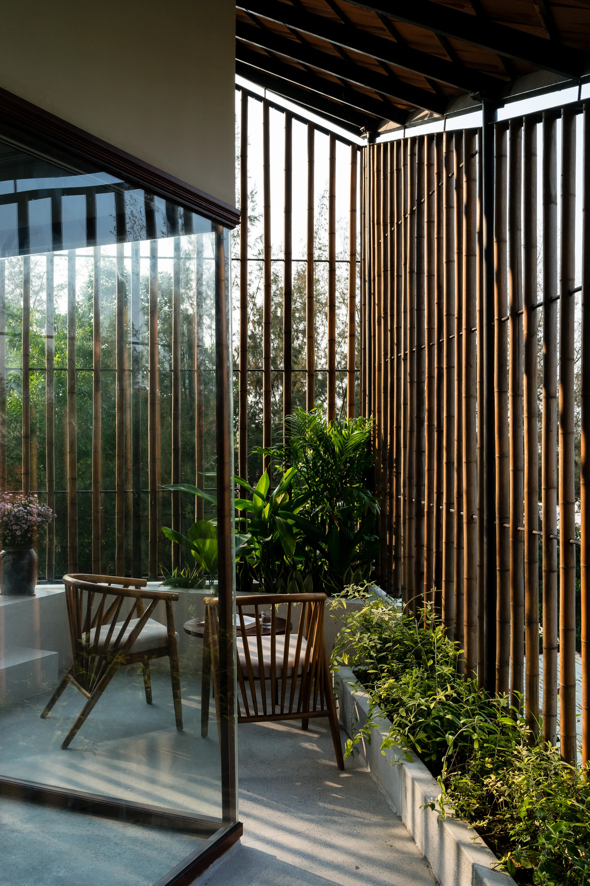 A covered patio features wooden spindle armchairs, a small table, concrete planters with greenery, and a tall vertical bamboo privacy screen.