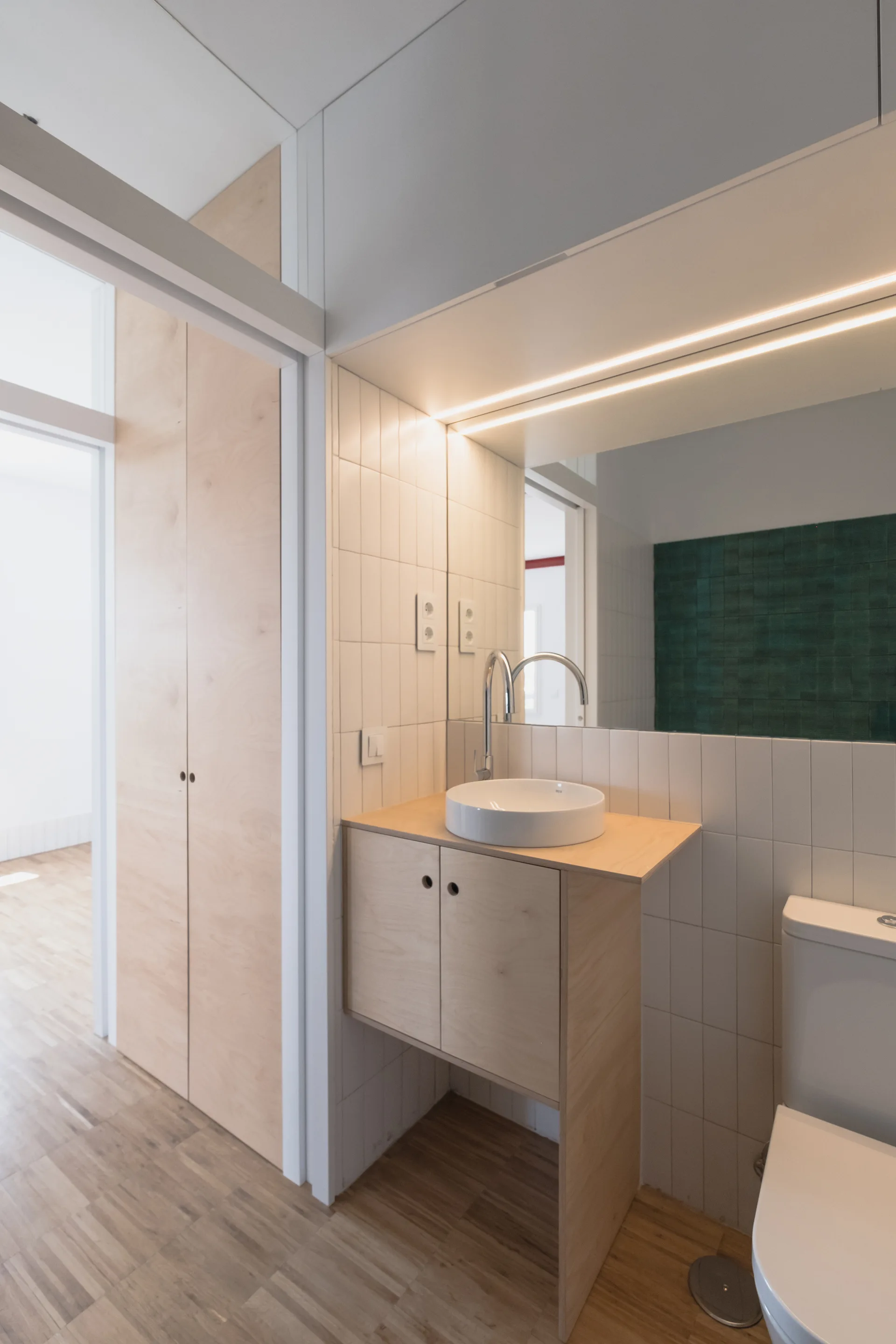A minimalist bathroom featuring a light wood vanity with a white vessel sink, vertical white subway tiles, a large mirror, and a white toilet.