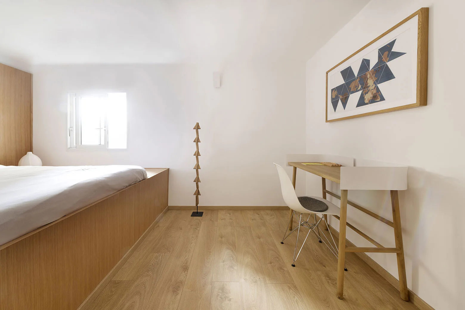 A minimalist bedroom with a light wood platform bed, a slim wooden desk with a white chair, and a geometric sculpture lamp against white walls.