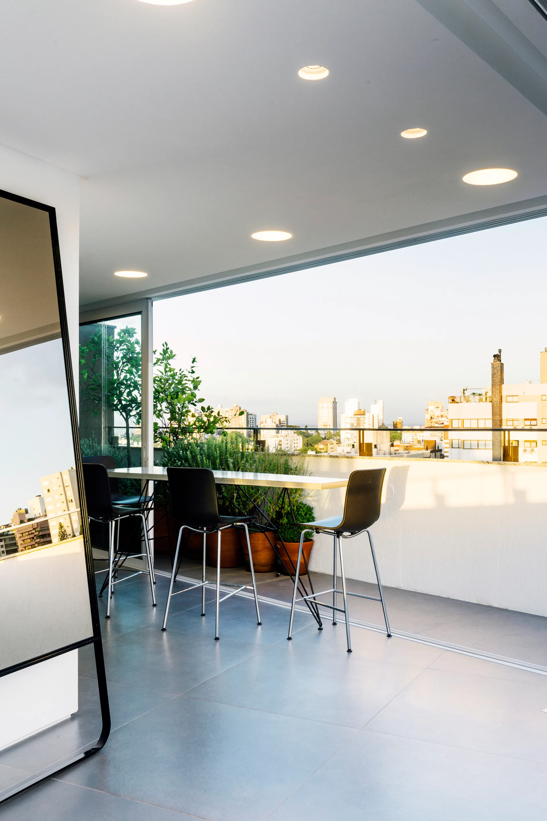 A modern apartment balcony features a tall black dining set, large format grey floor tiles, and potted green plants against a city skyline backdrop.