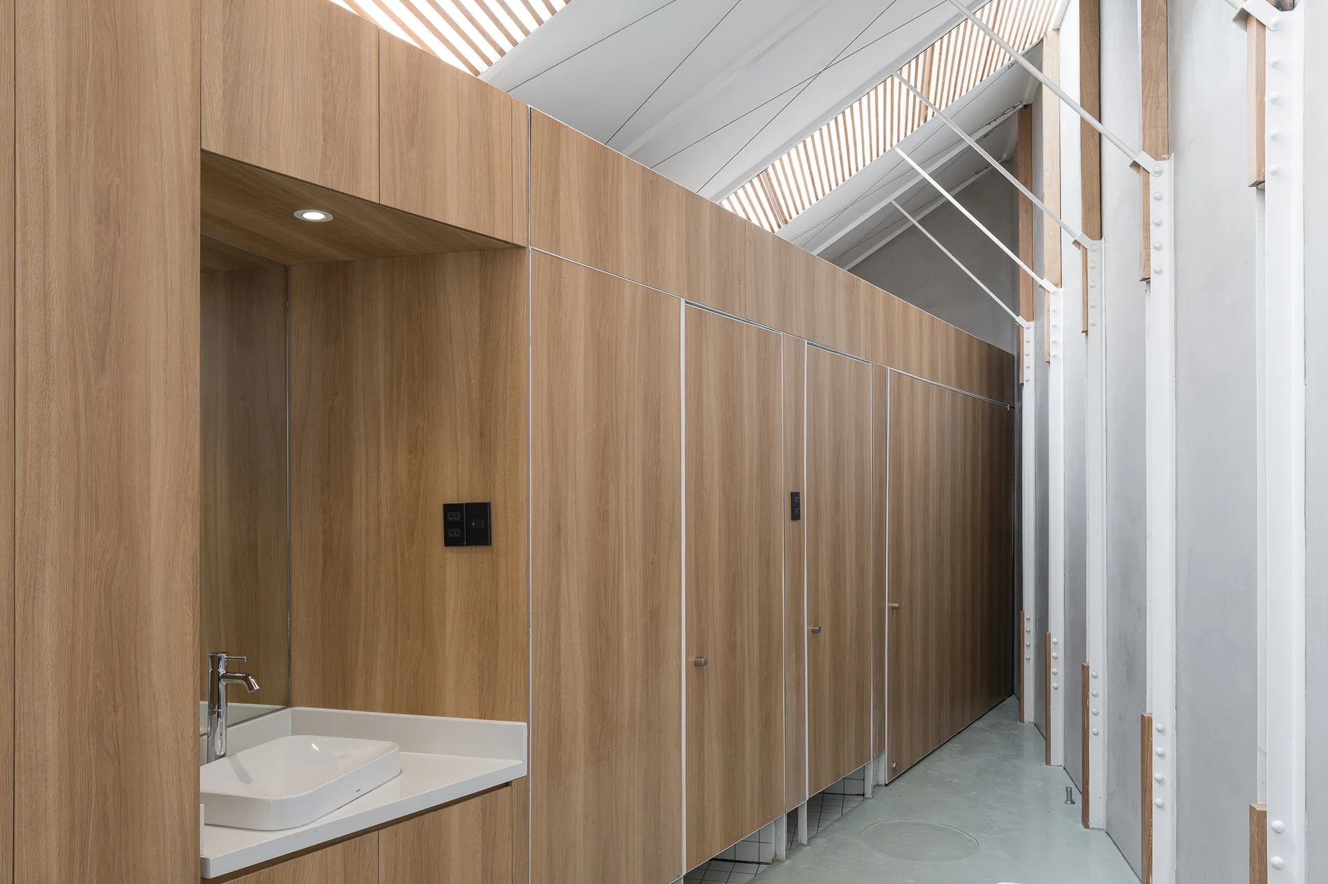 A modern restroom with light wood-grained stall partitions, a recessed white ceramic sink, and a slanted ceiling with skylight slats.
