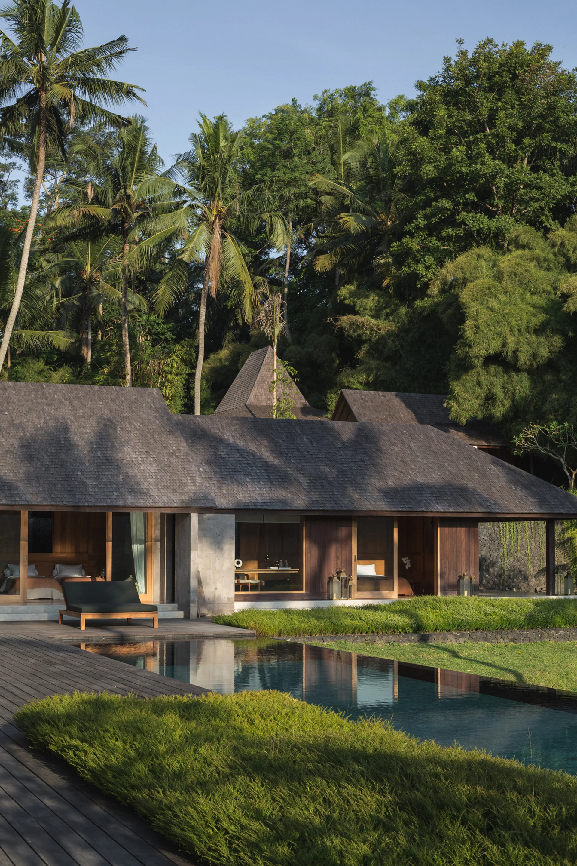 A dark-stained wooden villa with a shingle roof faces a swimming pool and wooden deck surrounded by tropical trees.