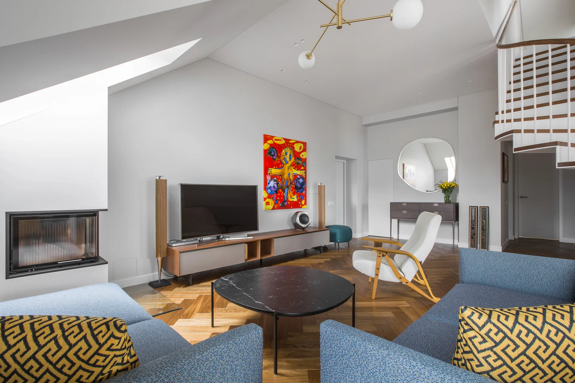 A contemporary attic living room features textured blue sofas, a round black marble coffee table, and a curved staircase under a sloped ceiling.