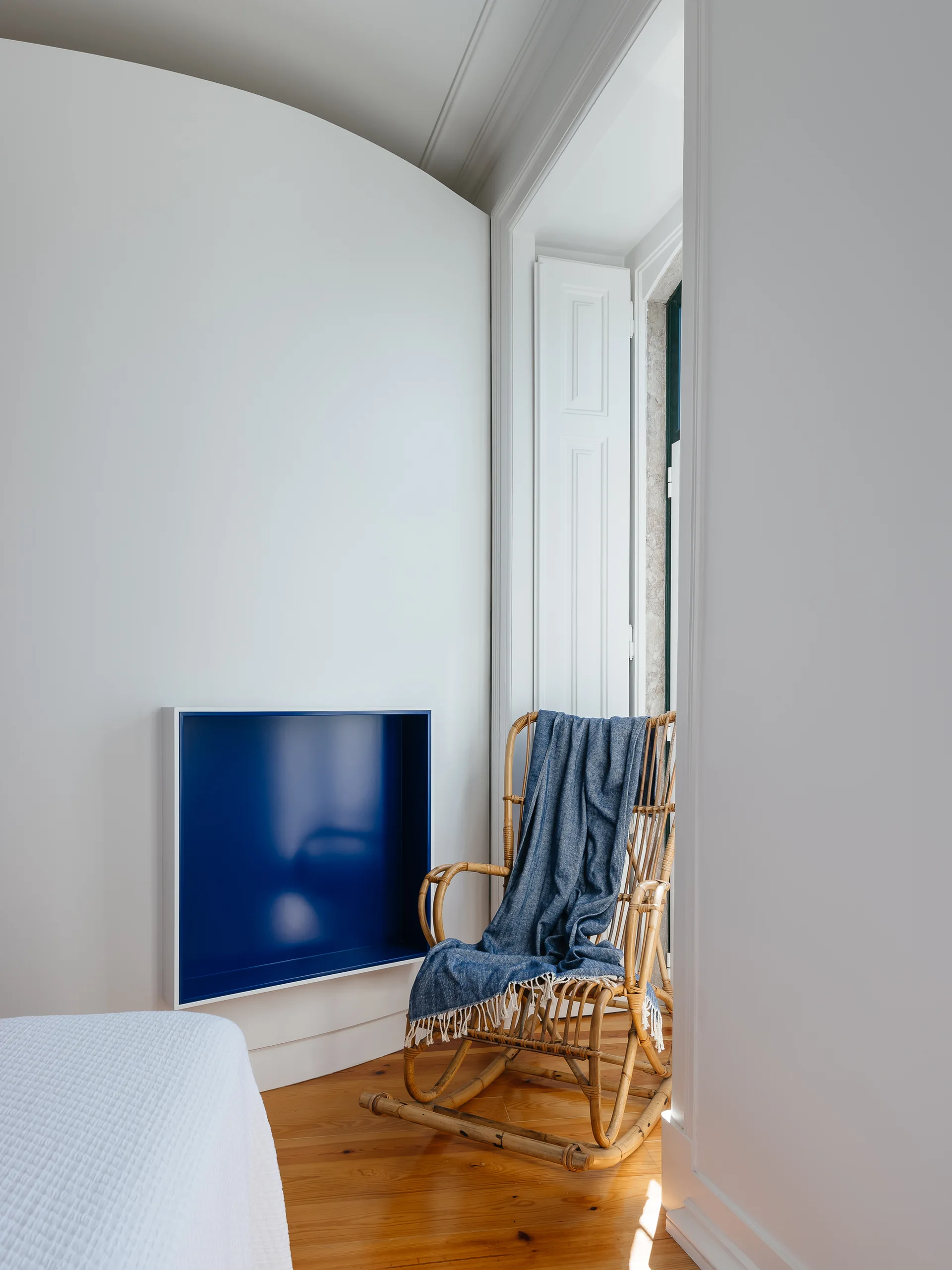 A bedroom corner features a rattan rocking chair with a blue blanket next to a white curved wall containing a blue recessed shelf.