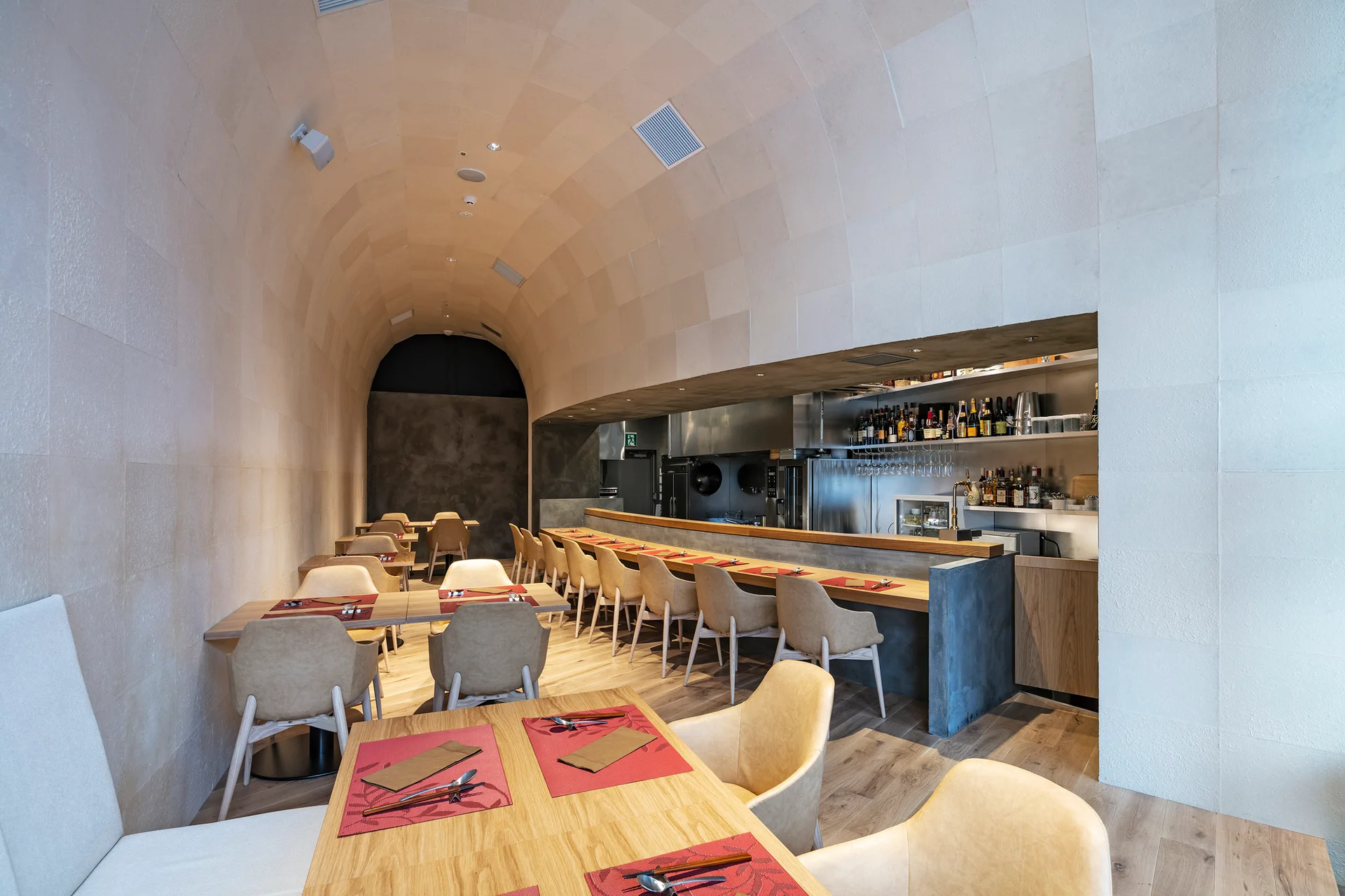 A dining space features a high barrel-vaulted ceiling tiled in beige stone, light wood tables with beige chairs, and a concrete bar.