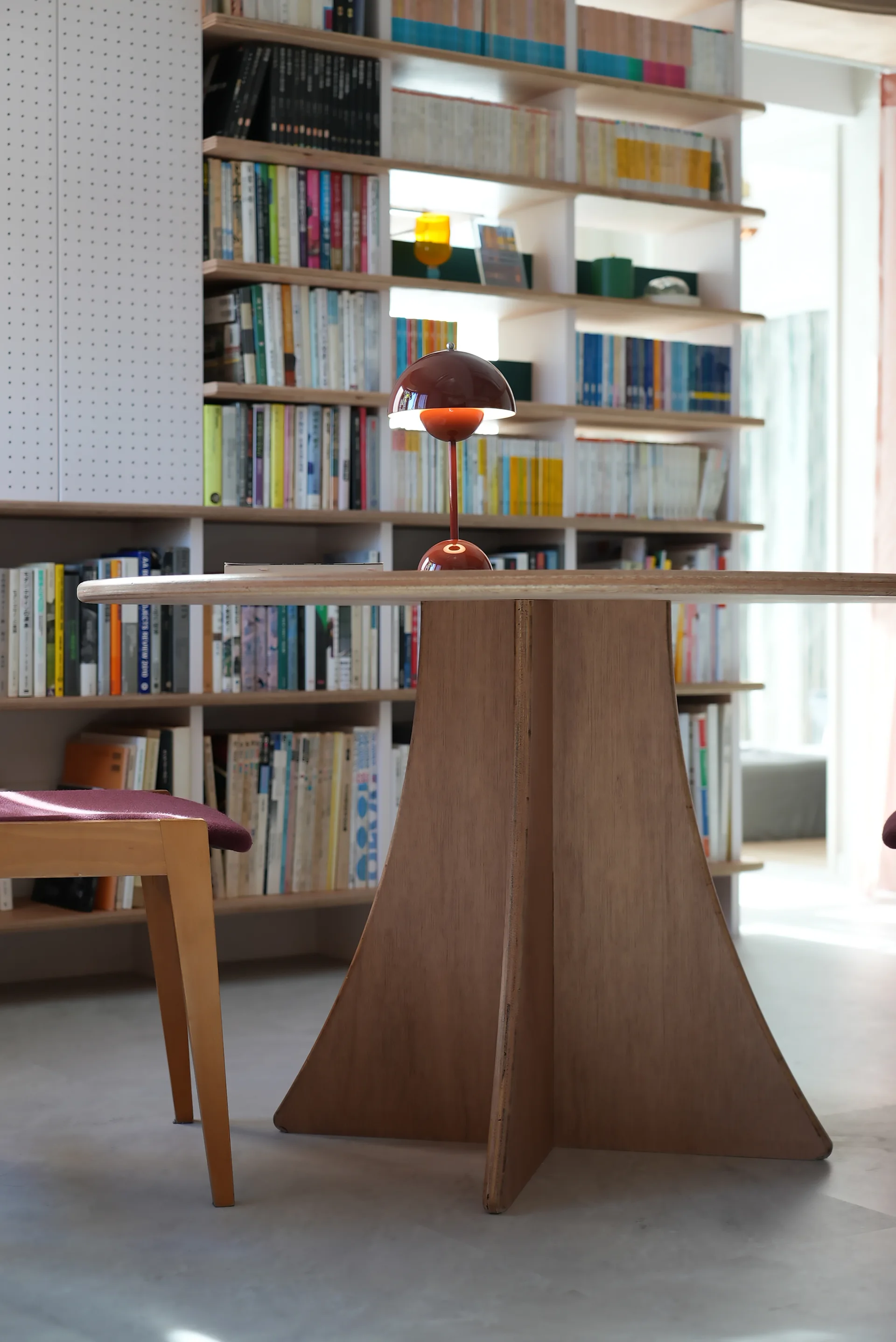 A small red dome lamp sits on a round wooden table in front of floor-to-ceiling bookshelves packed with books.