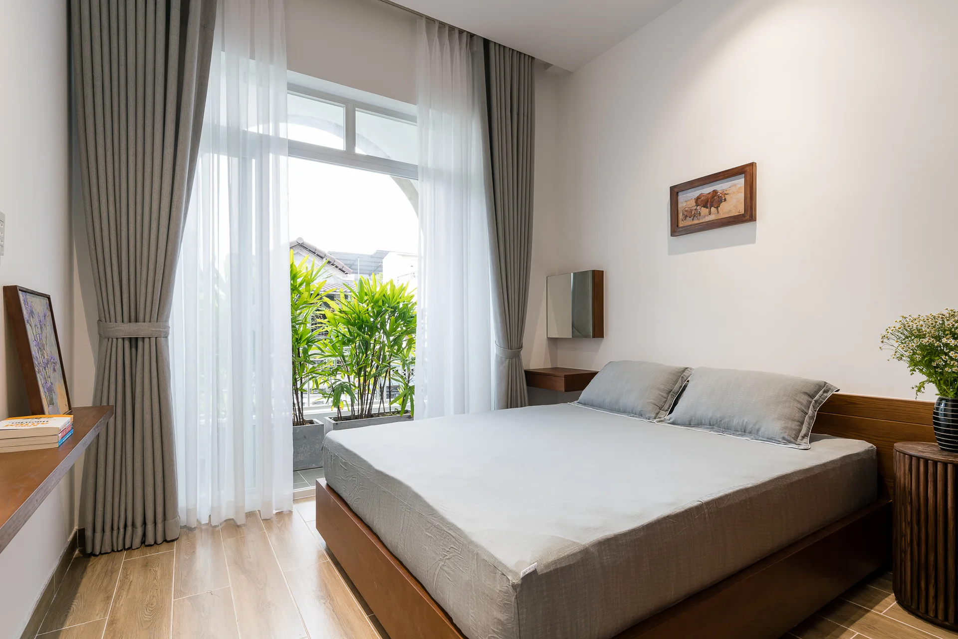 A modern bedroom features a wooden platform bed with grey sheets, a fluted wooden nightstand, and large windows with sheer white and grey drapes.