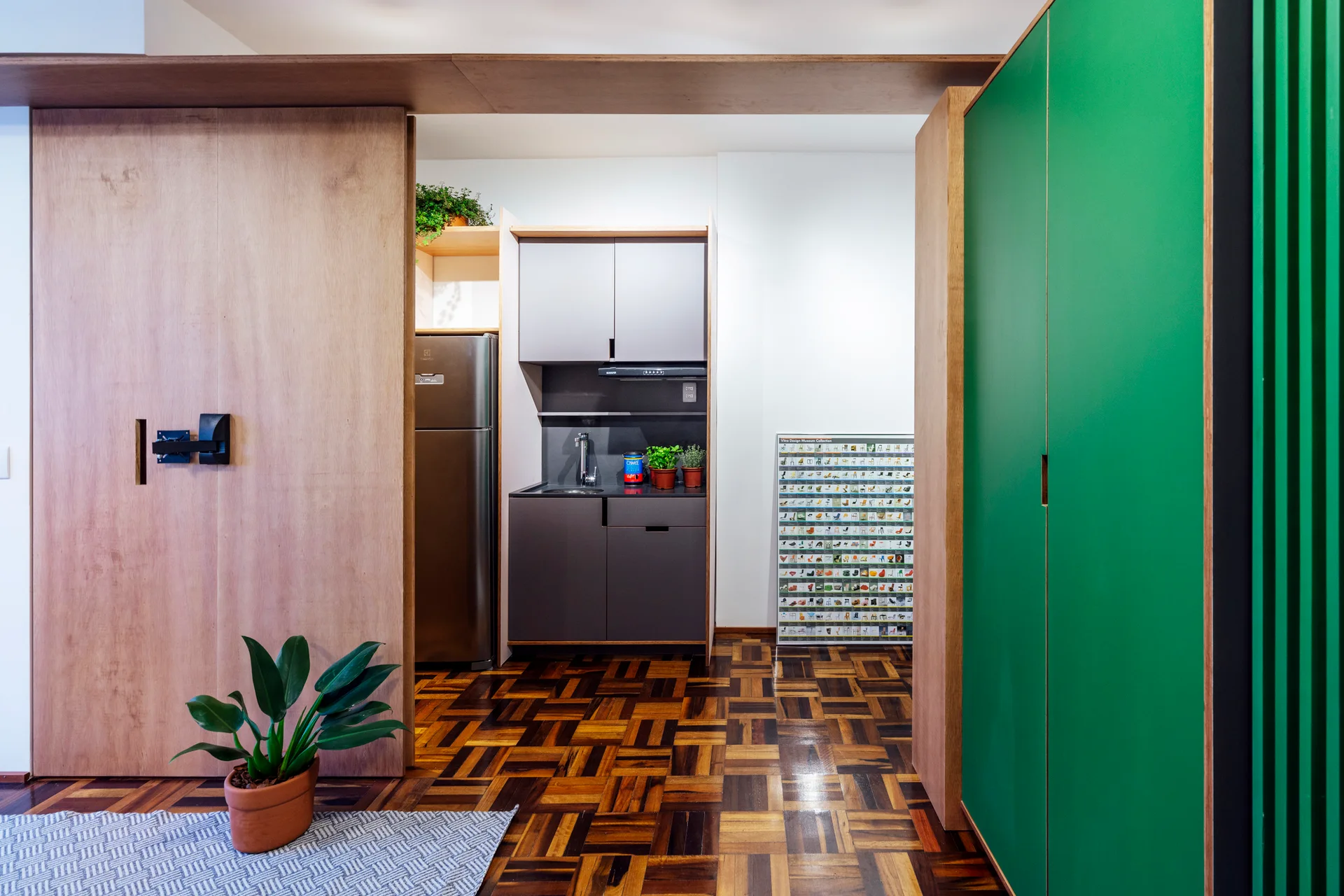 A compact grey kitchenette is flanked by warm wood panels and a vibrant green cabinet over geometric parquet wood flooring.