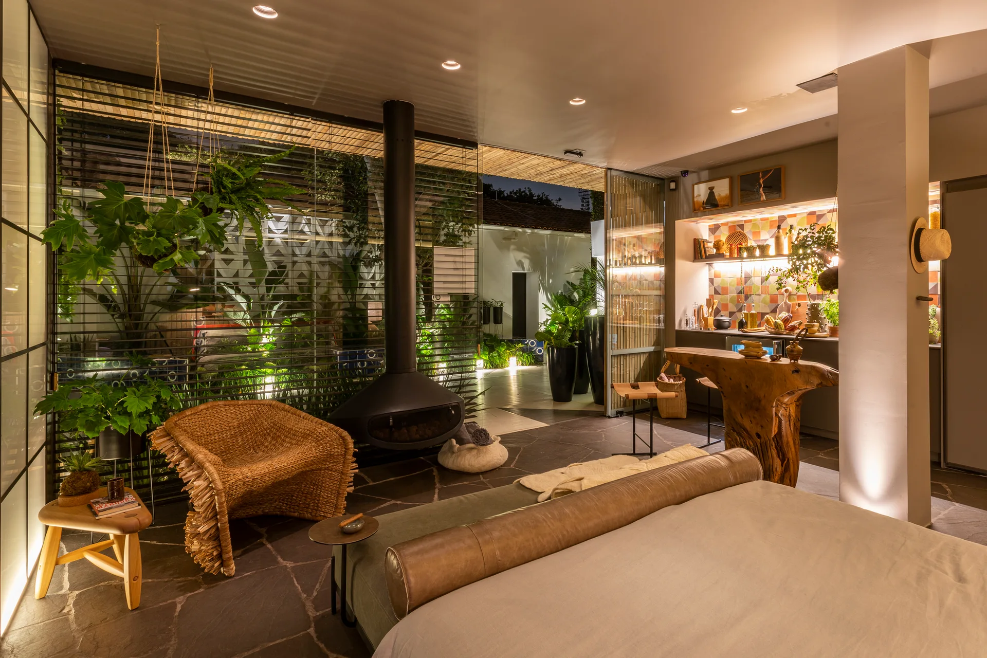 An open-plan bedroom features a suspended black fireplace, a woven armchair, a live-edge wood counter, and large windows looking out to a garden.