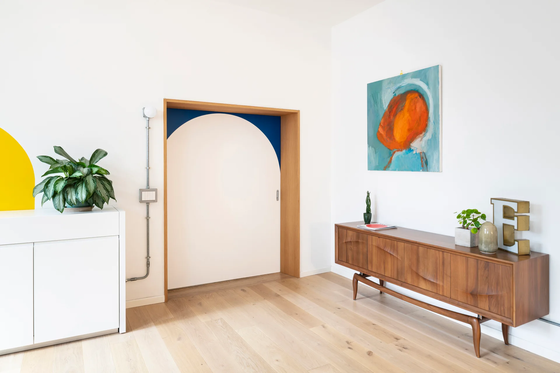 A bright white room features a mid-century wooden sideboard, an abstract blue painting, and a unique wood-trimmed doorway with a painted arch.